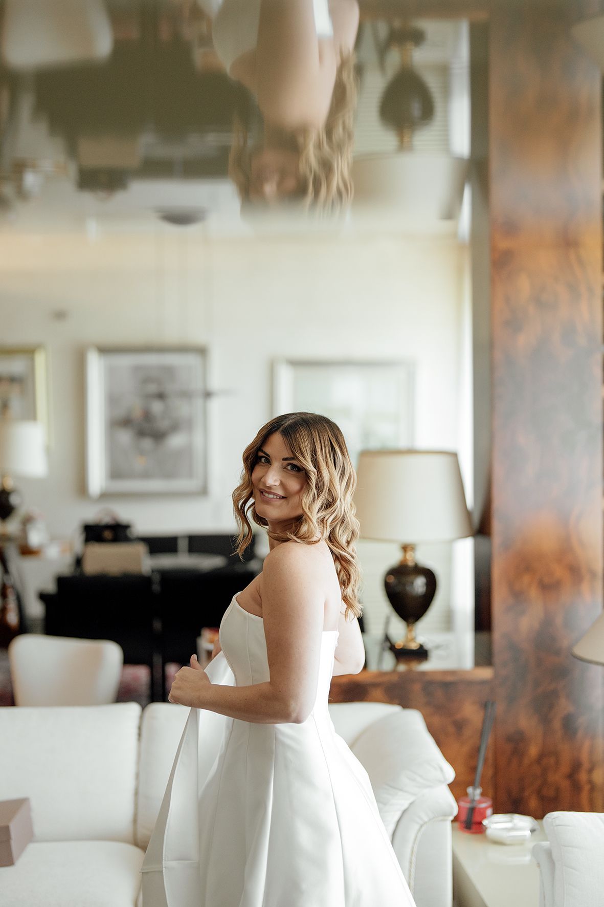 A woman in an elegant white wedding dress stands and poses in an upscale interior room with modern decor.