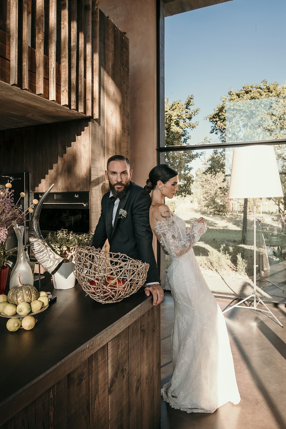 Couple in formal wedding attire stand at wooden counter with decor and natural lighting streaming through windows.