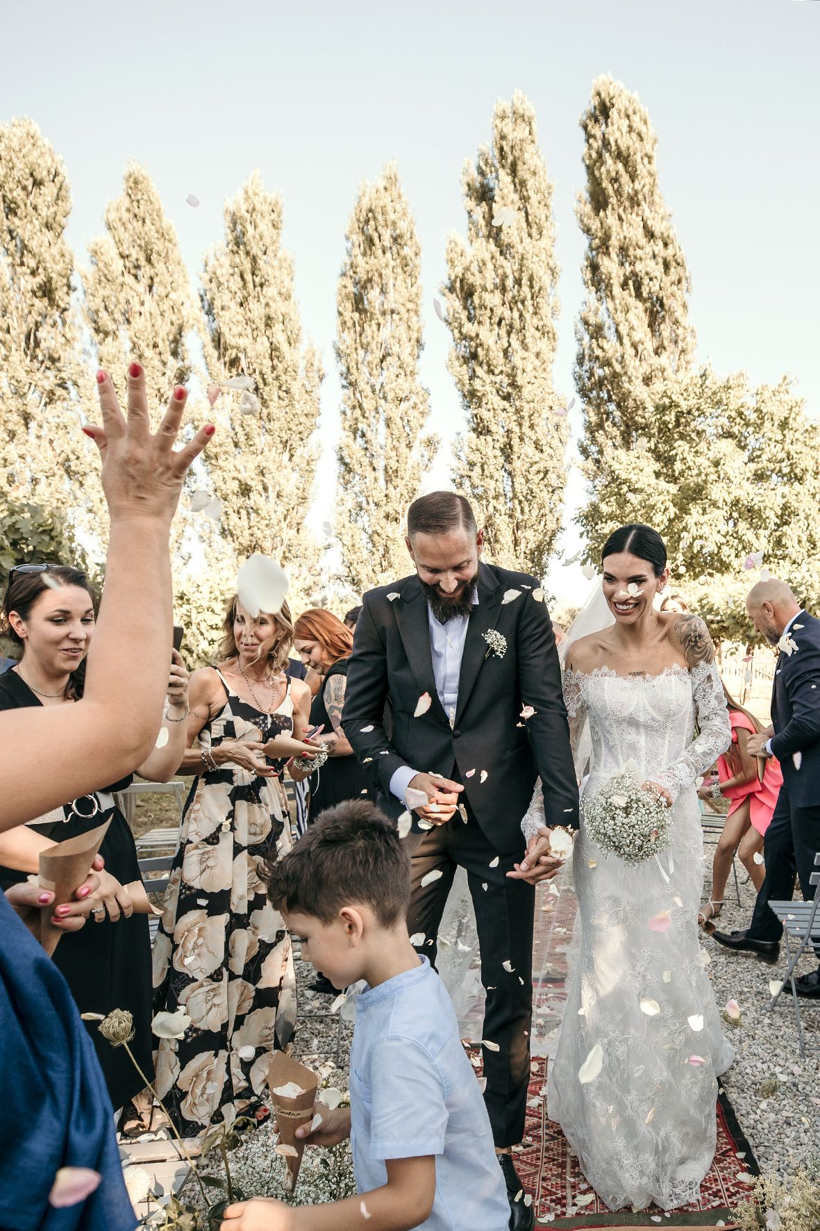 Wedding guests celebrate with confetti toss in front of tall cypress trees on a sunny afternoon.