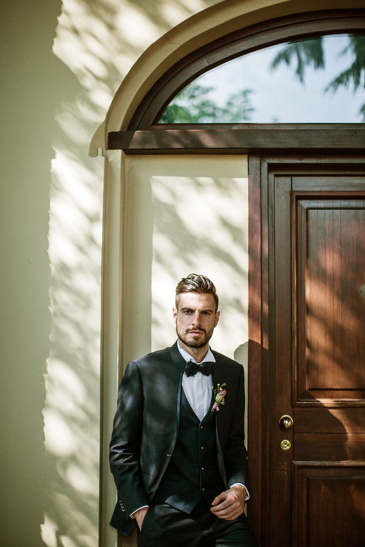 A well-dressed person in a classic black tuxedo with bow tie stands elegantly by a wooden door in natural lighting.