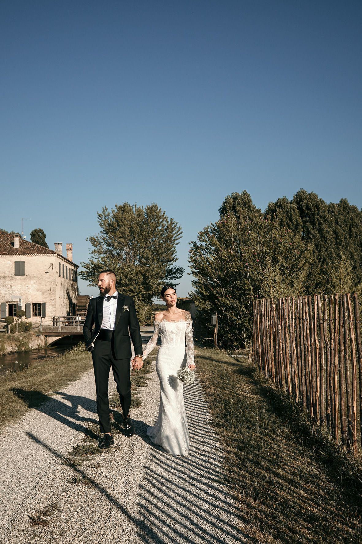 Couple in formal attire walks along a gravel path beside a rustic stone house and wooden fence on a sunny day.