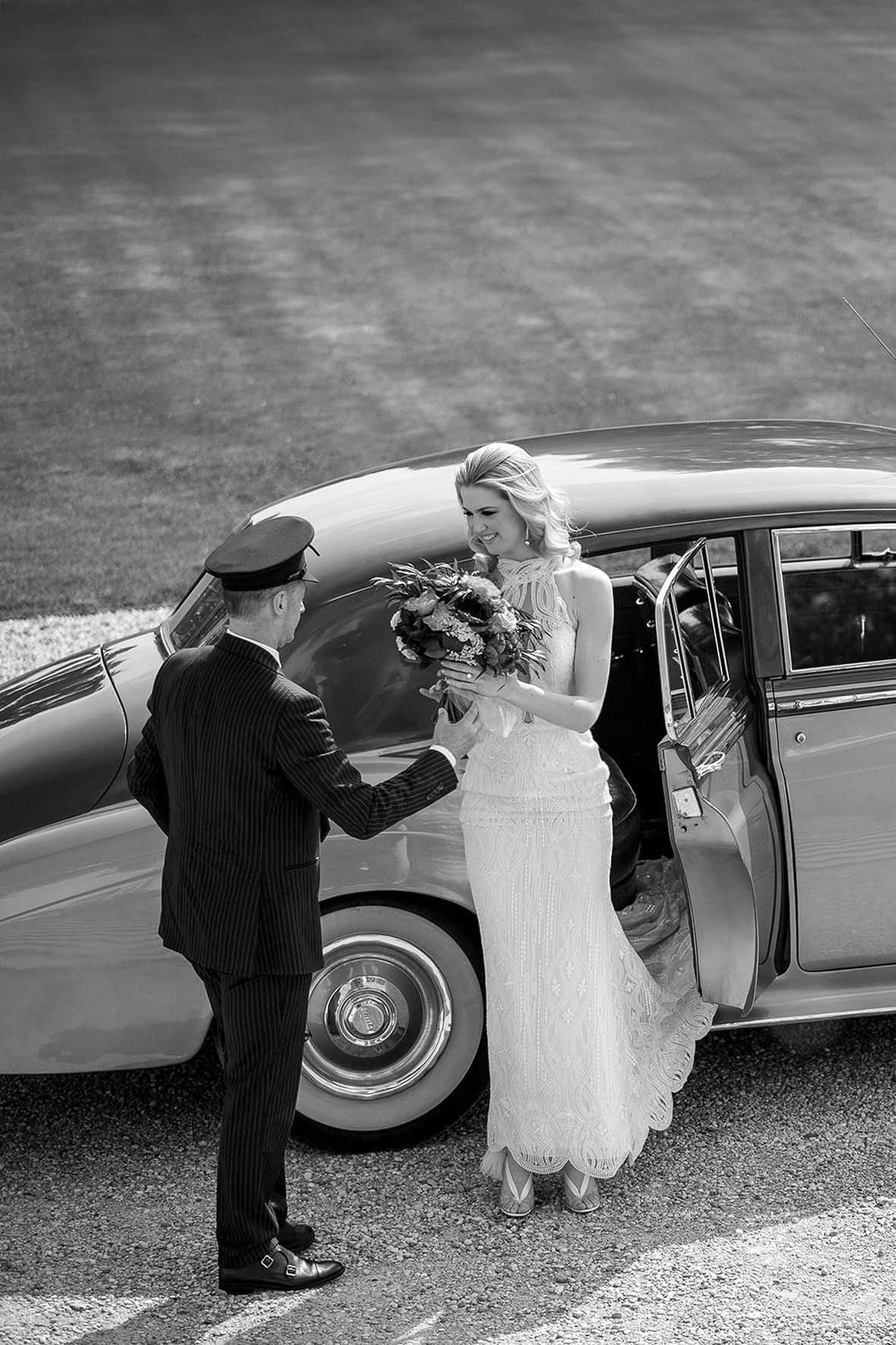 A chauffeur helps someone in a white wedding dress exit a vintage luxury car in black and white photography.