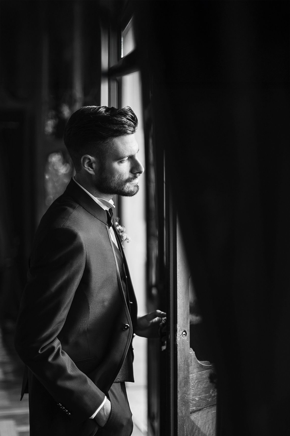 A black and white portrait of someone in a suit standing by a window in dramatic lighting.