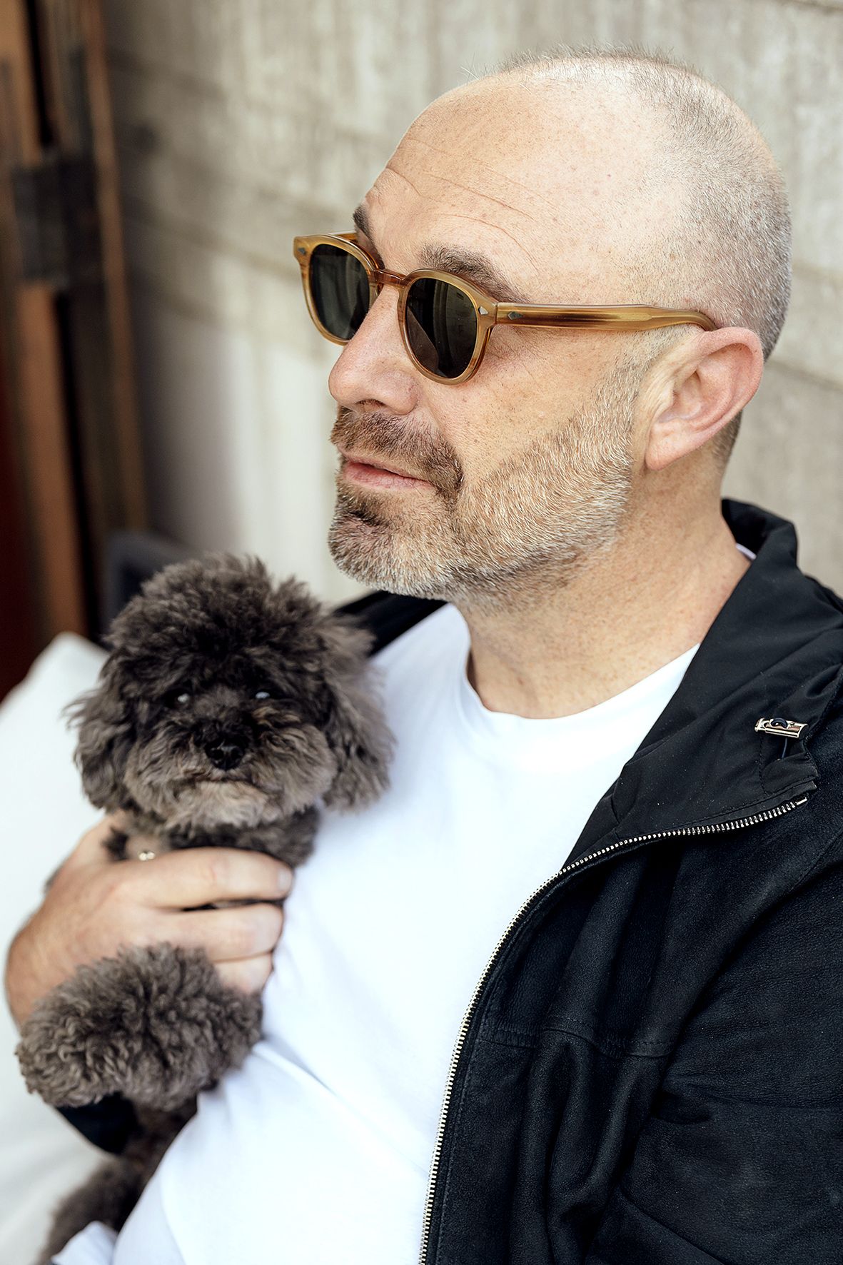 Person in sunglasses and black jacket holding a small fluffy grey dog while standing against a wall.