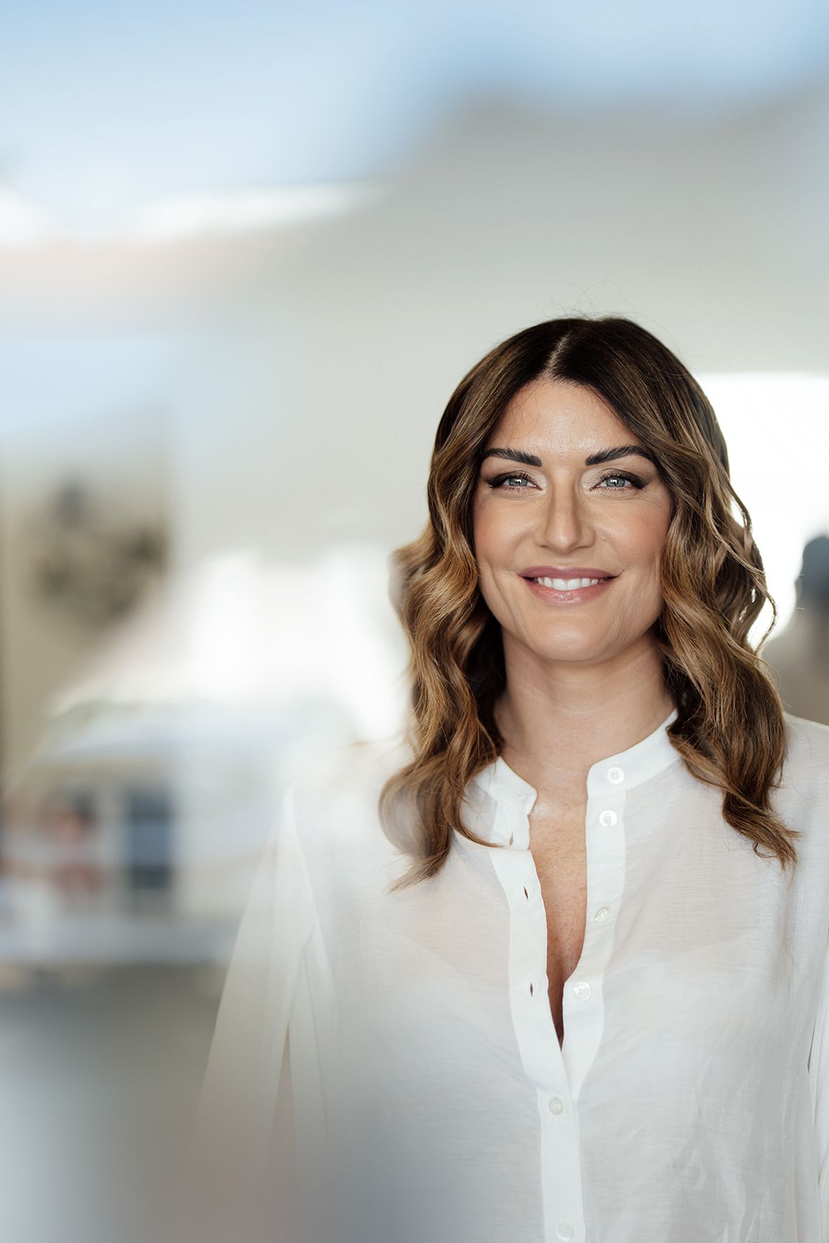 Professional portrait showing a person with wavy brown hair wearing a white button-up blouse against a blurred background.