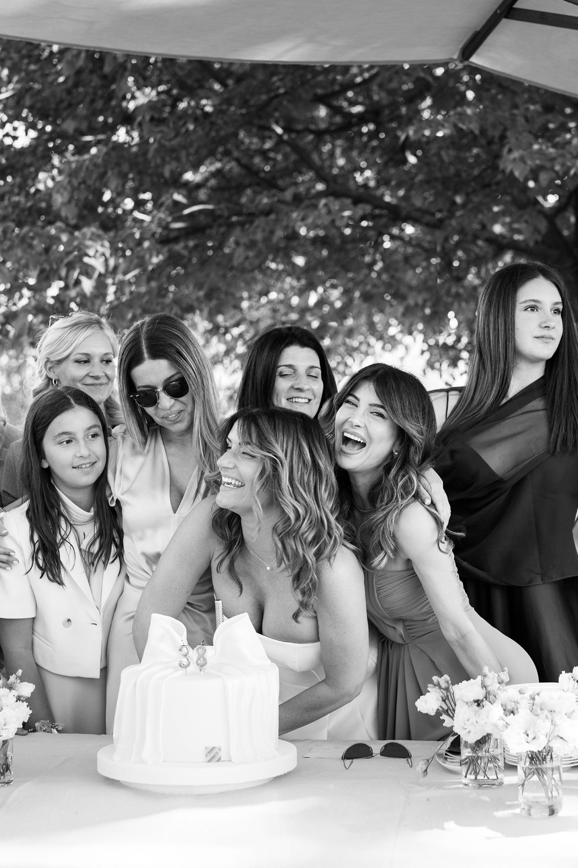 Black and white photo of a group of friends gathered around a simple white birthday cake at an outdoor celebration.