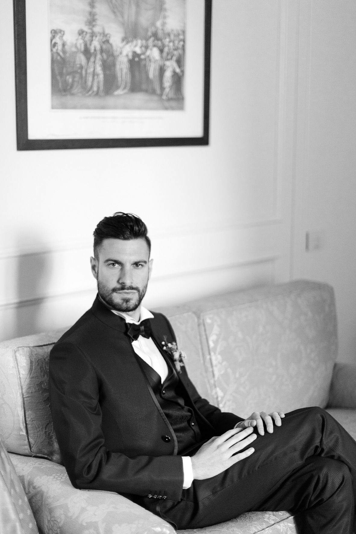 A black and white formal portrait of someone in a tuxedo with bow tie sitting elegantly on a couch.