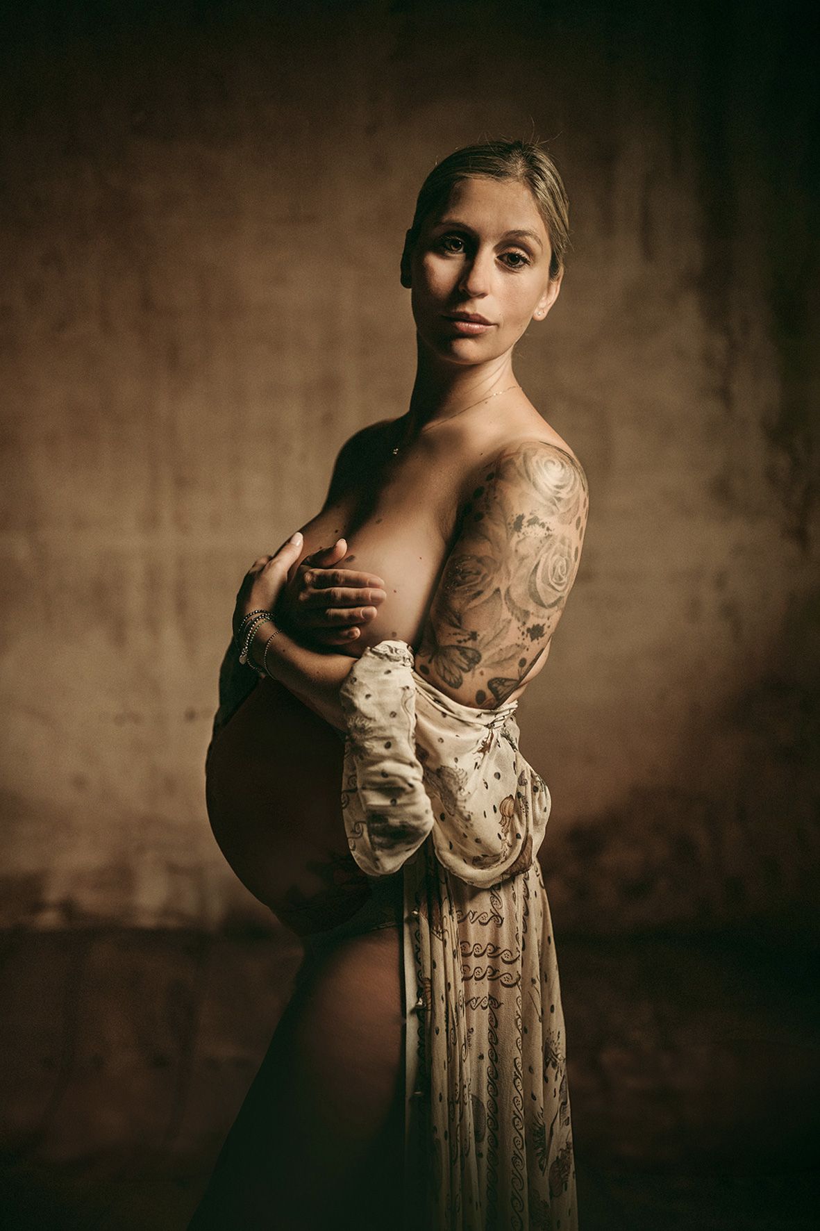 Artistic maternity portrait in sepia tones featuring an elegant lace dress and tattoo sleeve, photographed against textured backdrop.