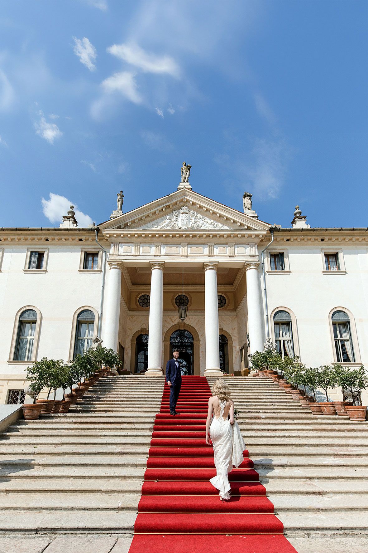 Grand neoclassical villa with white columns and red carpet staircase leading to ornate entrance under bright blue sky.