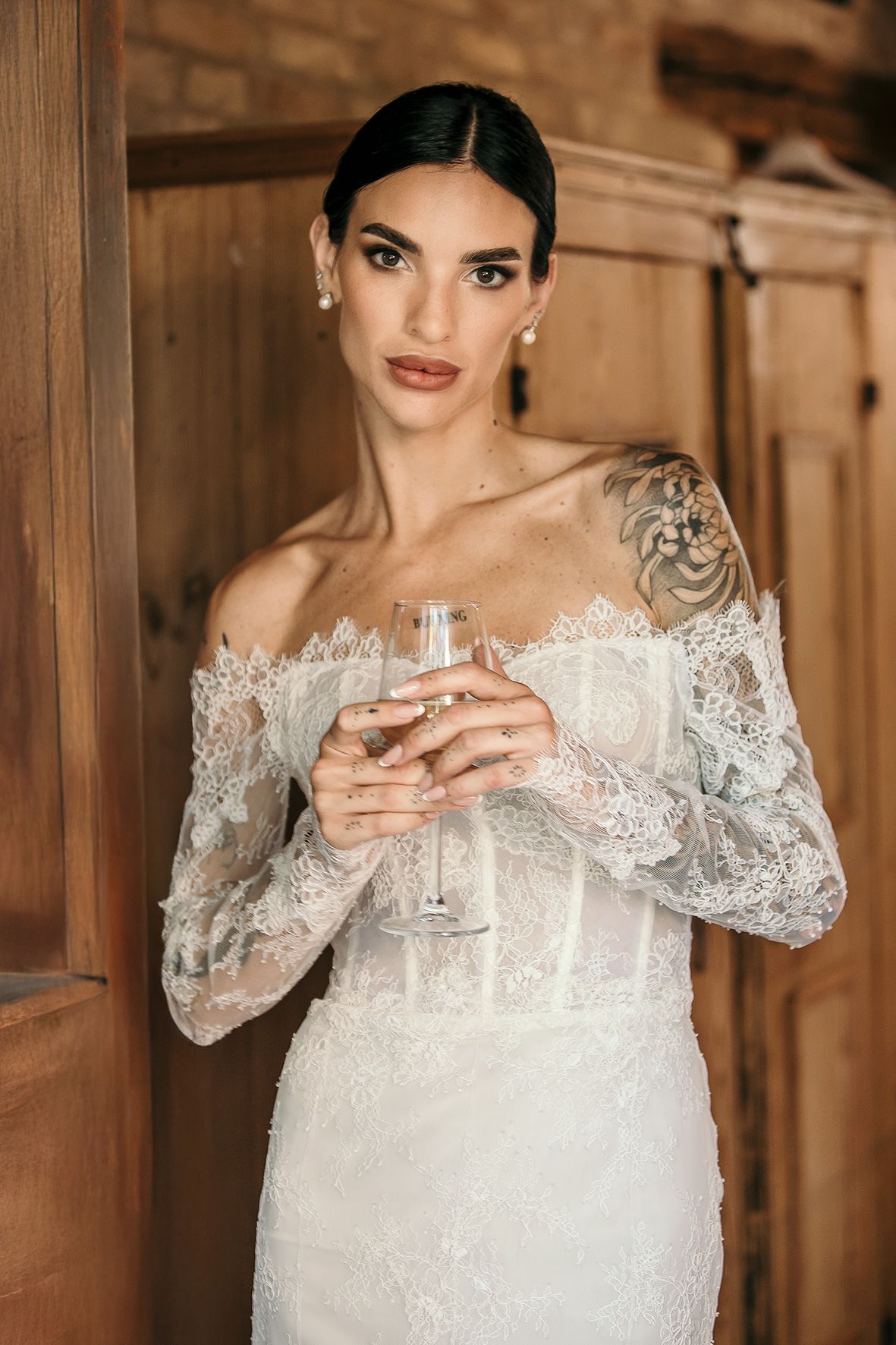 Elegant off-shoulder lace wedding dress with long sleeves shown in warm lighting while holding champagne glass.