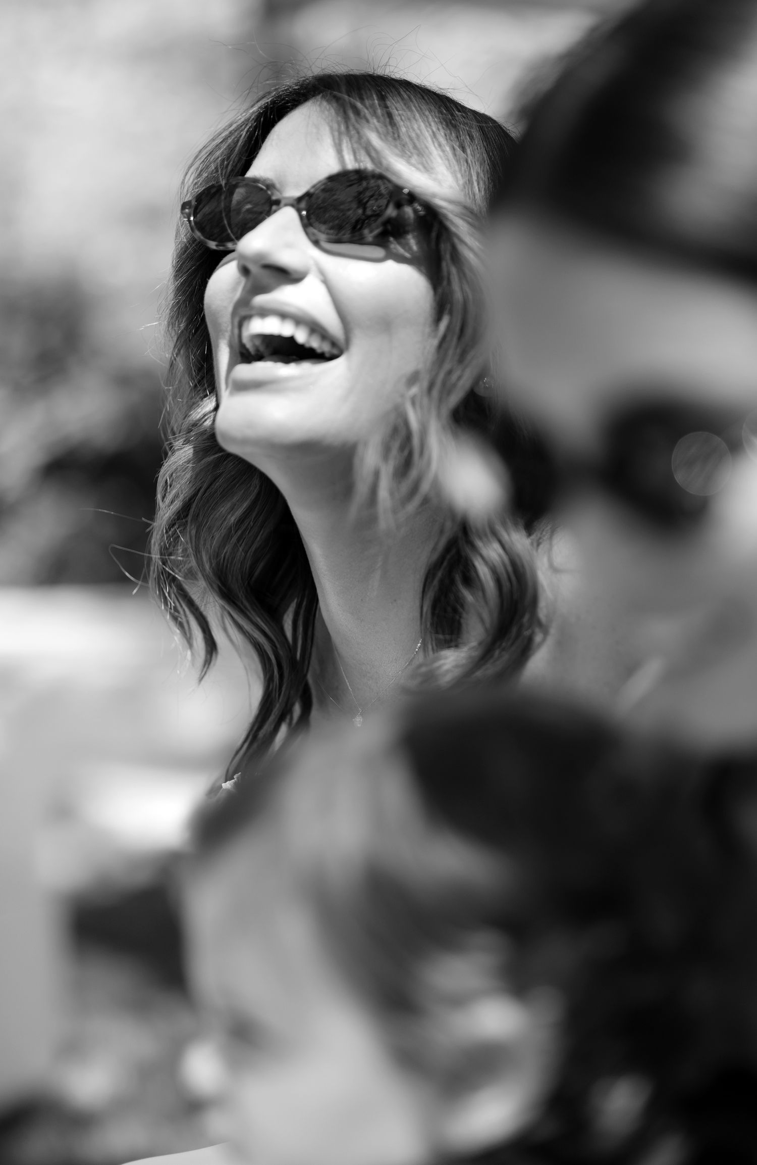 Black and white portrait of someone laughing joyfully while wearing sunglasses outdoors in natural light.