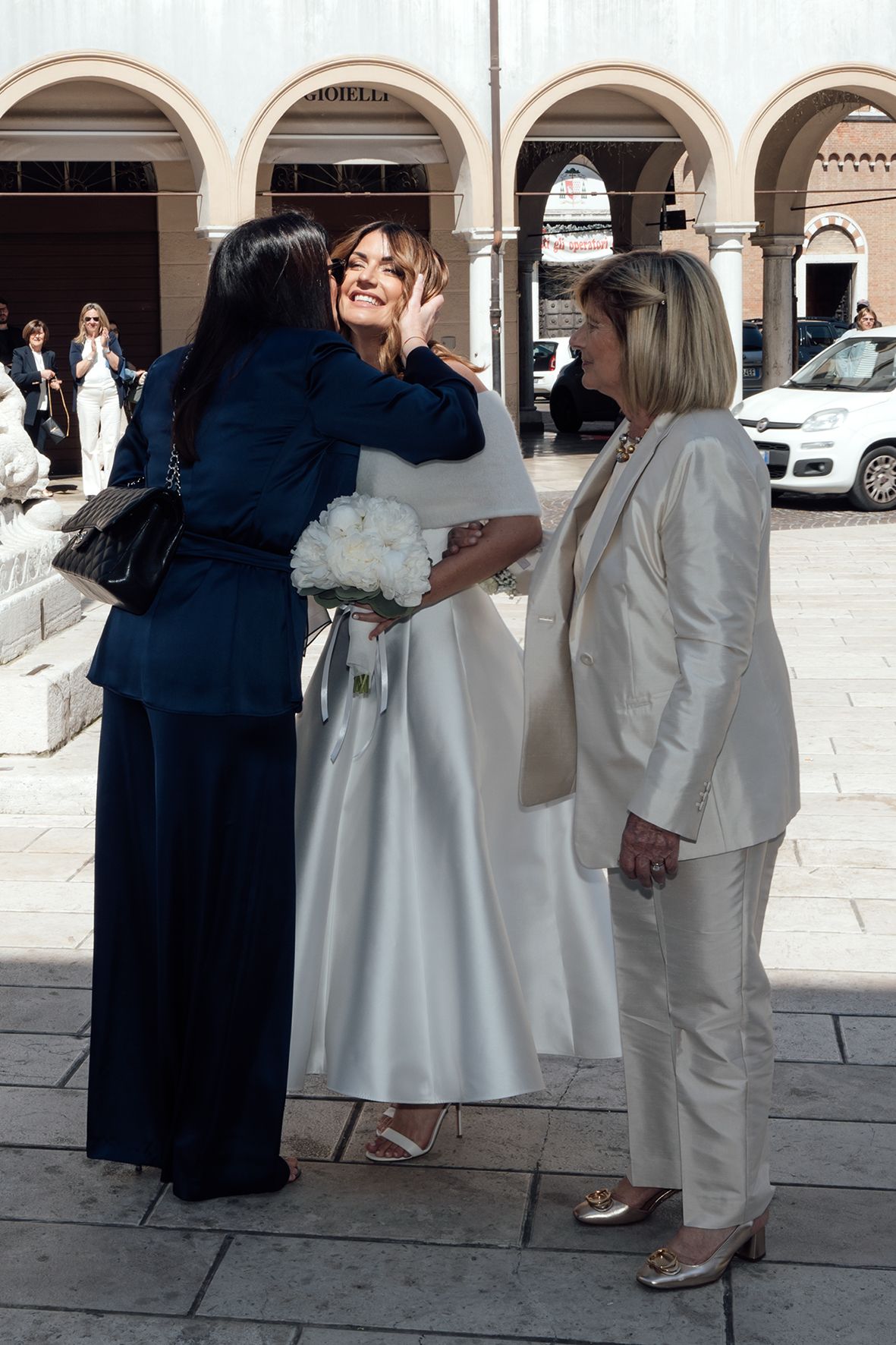 Three people sharing a joyful moment together under arched columns, with one wearing a white dress and others in dark and light outfits.