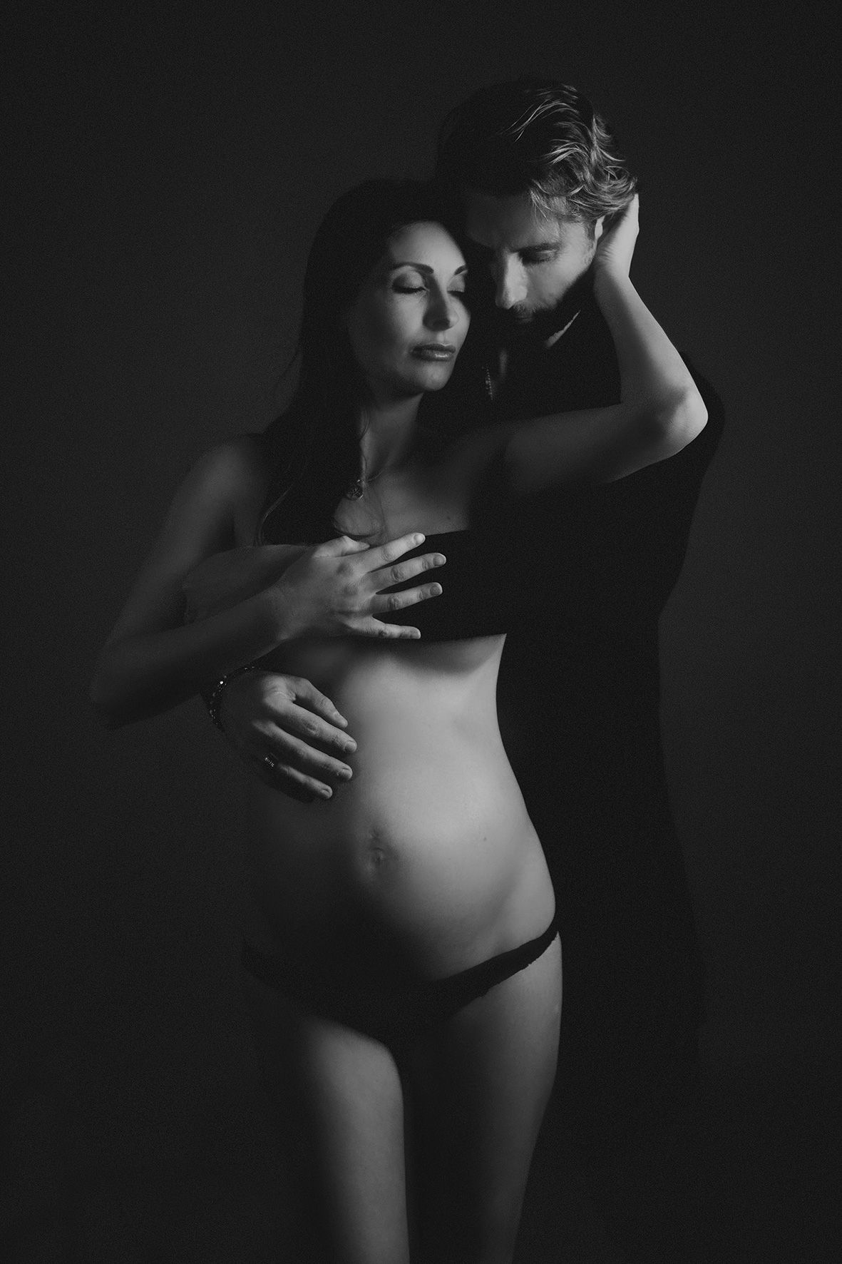 Black and white intimate maternity photo with dramatic lighting shows a couple embracing in an artistic pose.