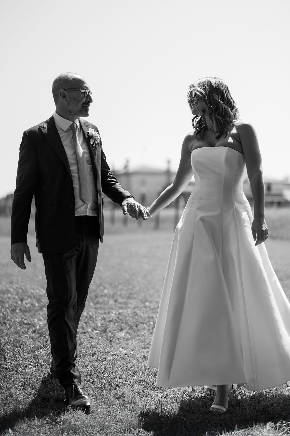 Black and white photo of a wedding couple walking hand in hand through a sunny meadow.