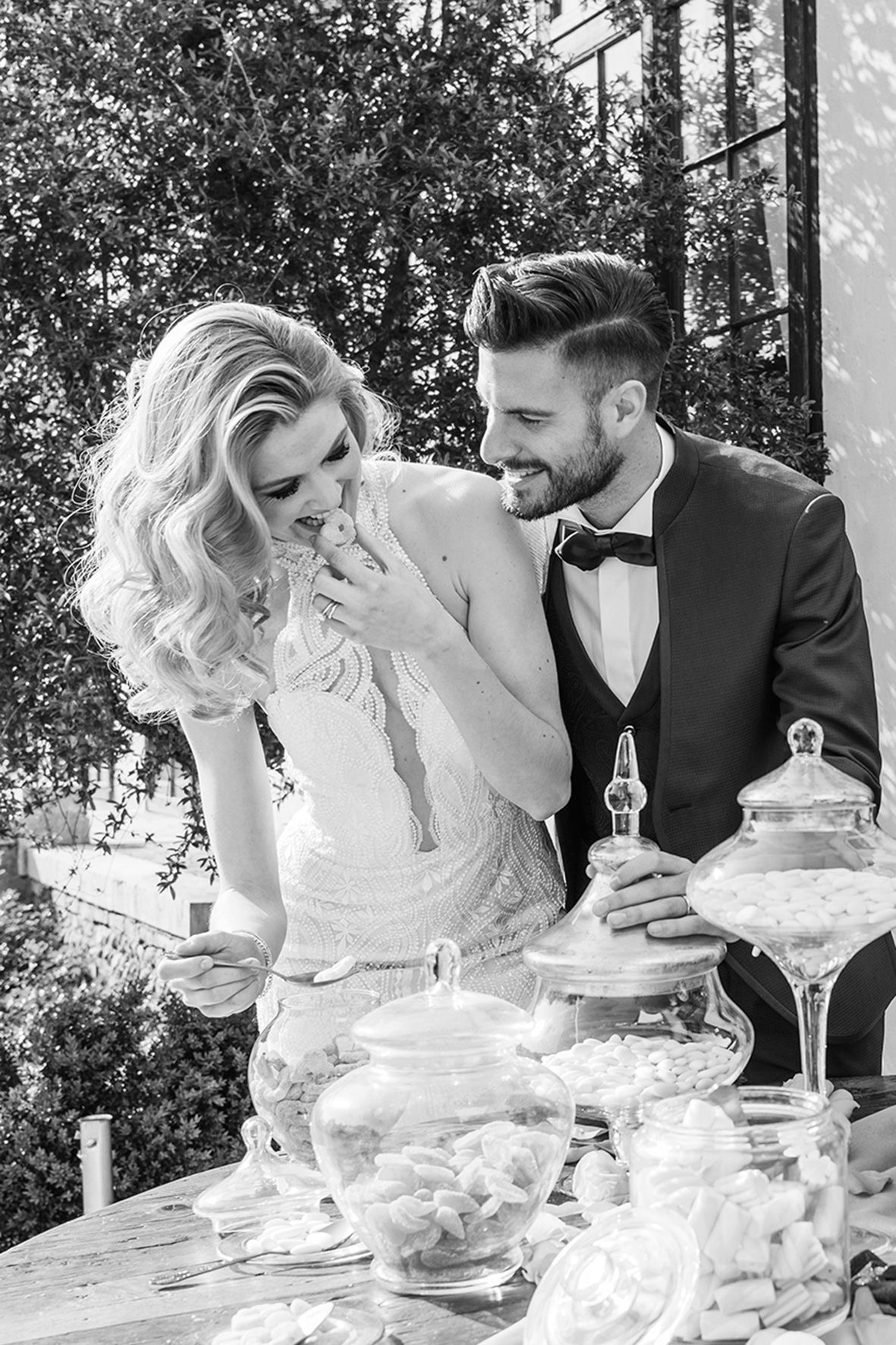 A joyful black and white wedding photo with a couple sharing a candid moment at a dessert table display outdoors.