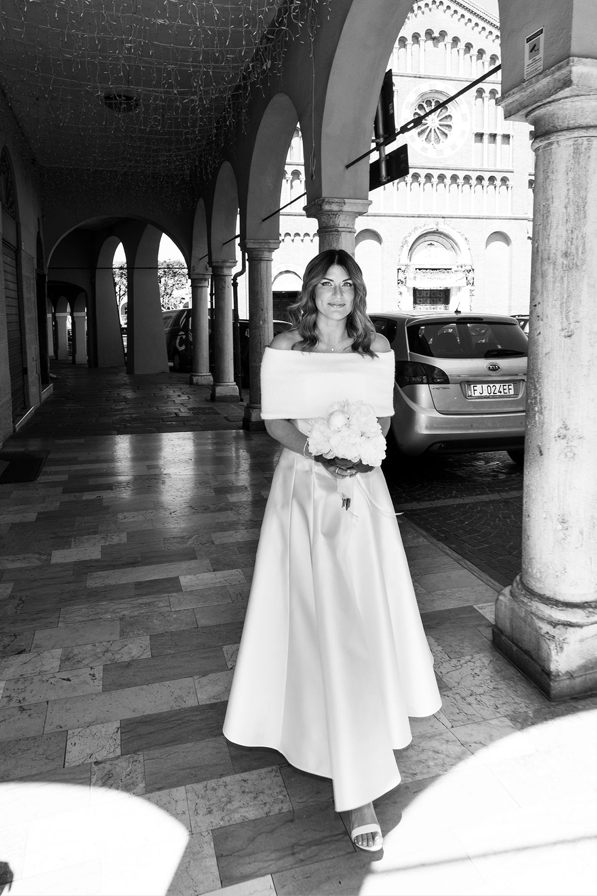 A wedding photo showing a bride in a white off-shoulder dress posing under a classical arched colonnade.