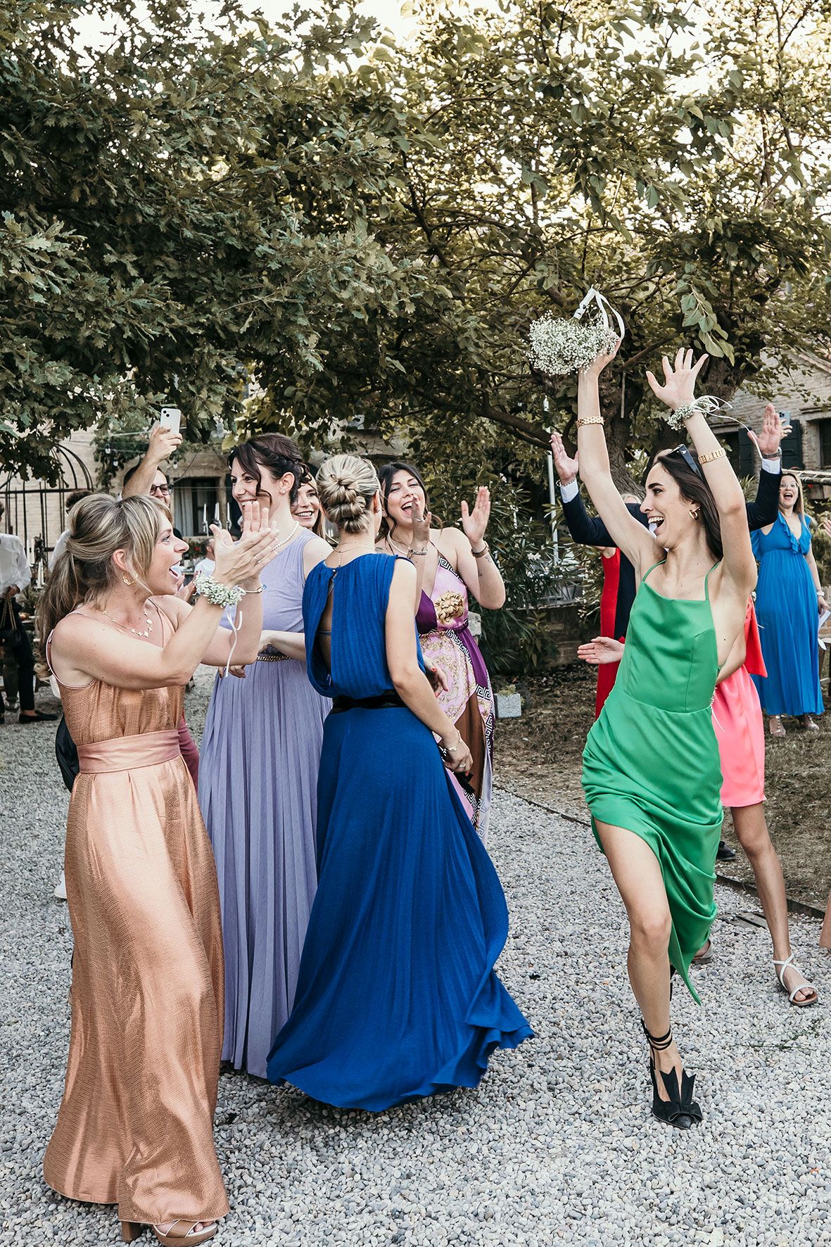 A group of wedding guests in colorful formal dresses excitedly reach for a bouquet toss at an outdoor celebration.