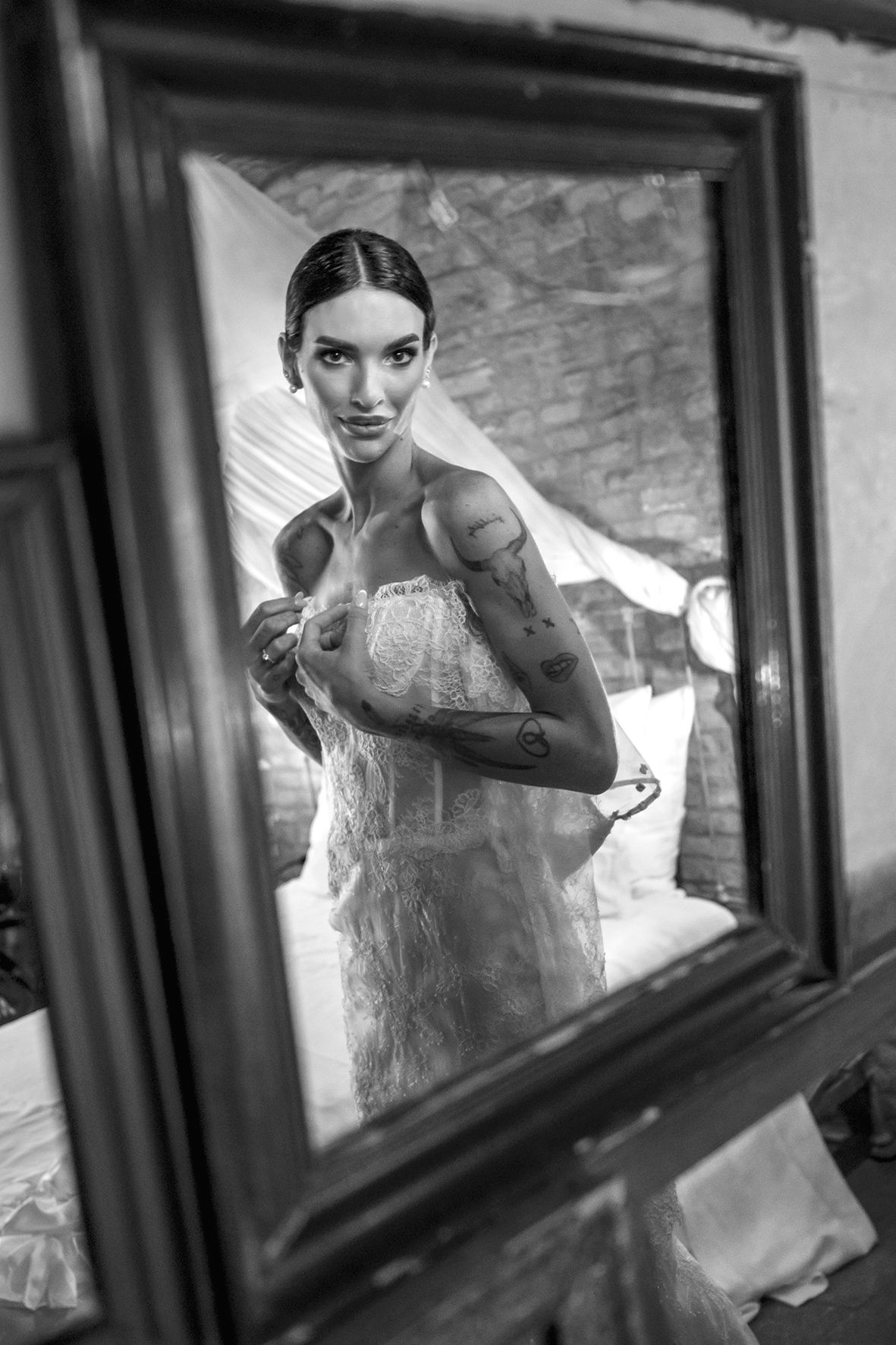 A black and white photograph of a bride getting ready, viewed through an ornate mirror frame.