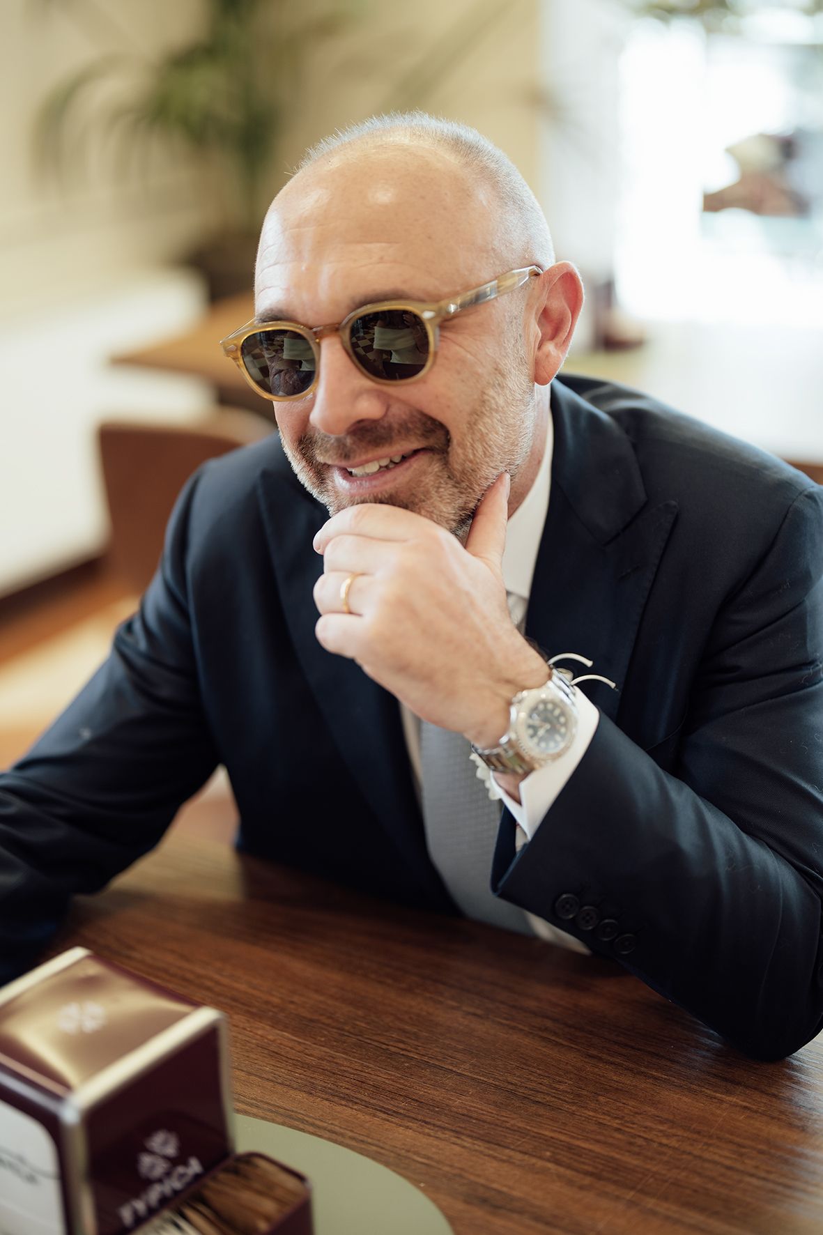 Stylish businessman in sunglasses and suit sits at wooden table wearing luxury watch with confident smile.