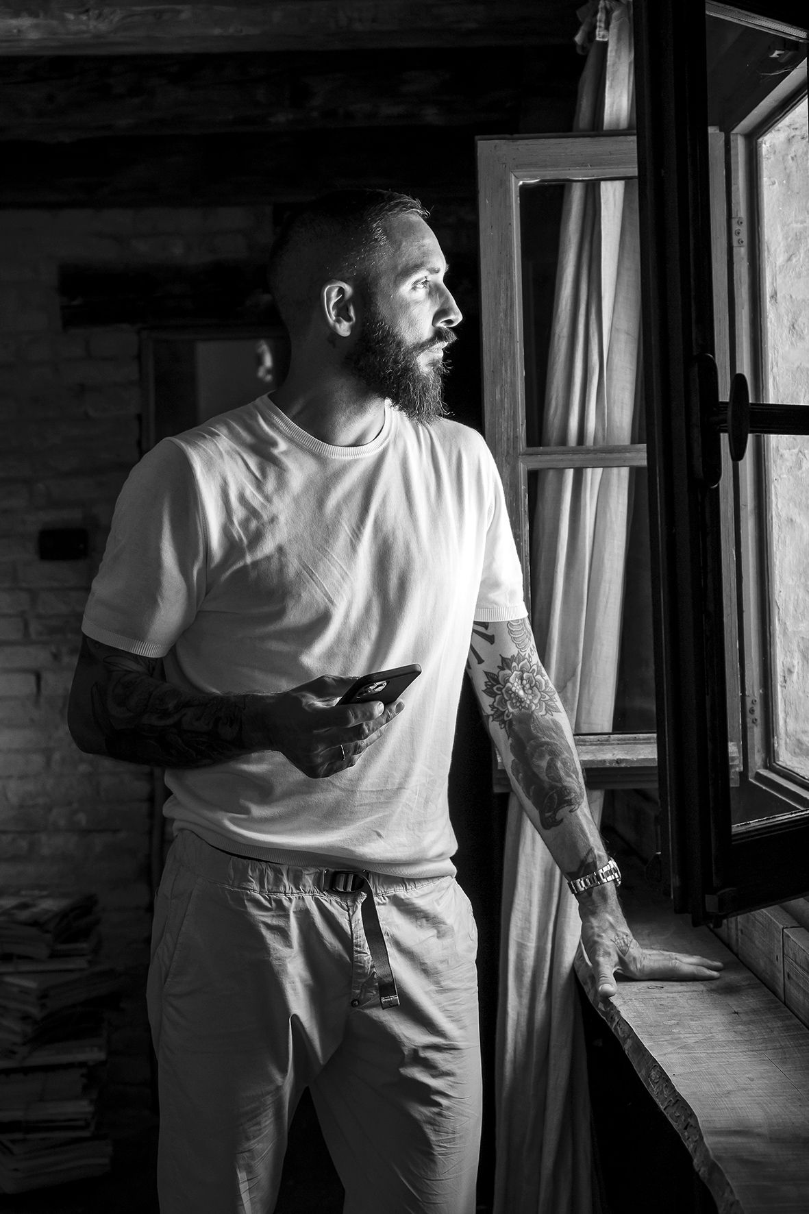 A monochrome photograph of someone in a white t-shirt and tattoos looking thoughtfully out a window in dramatic lighting.