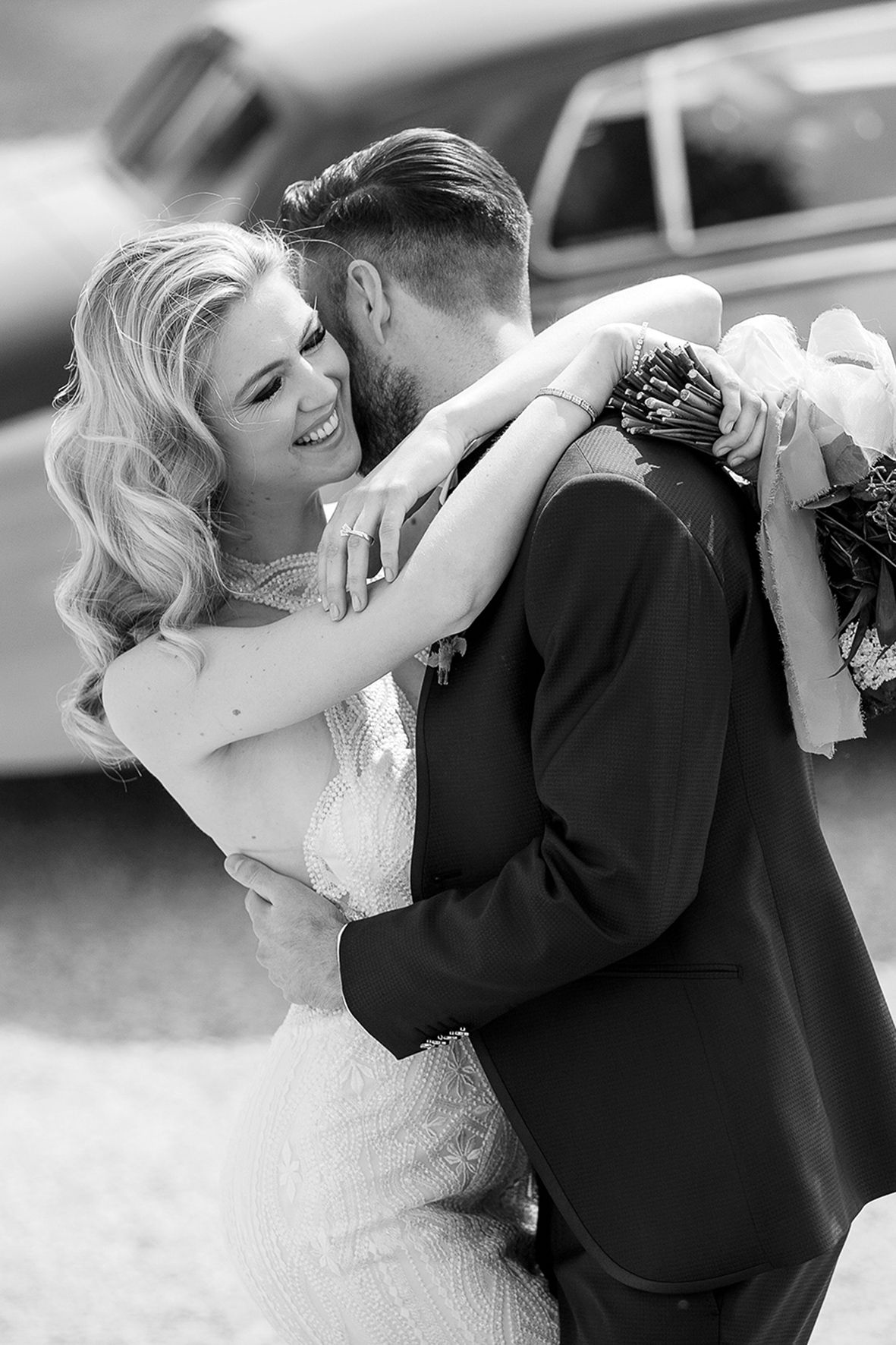 Black and white wedding photo of a romantic embrace between bride and groom near a vintage car.