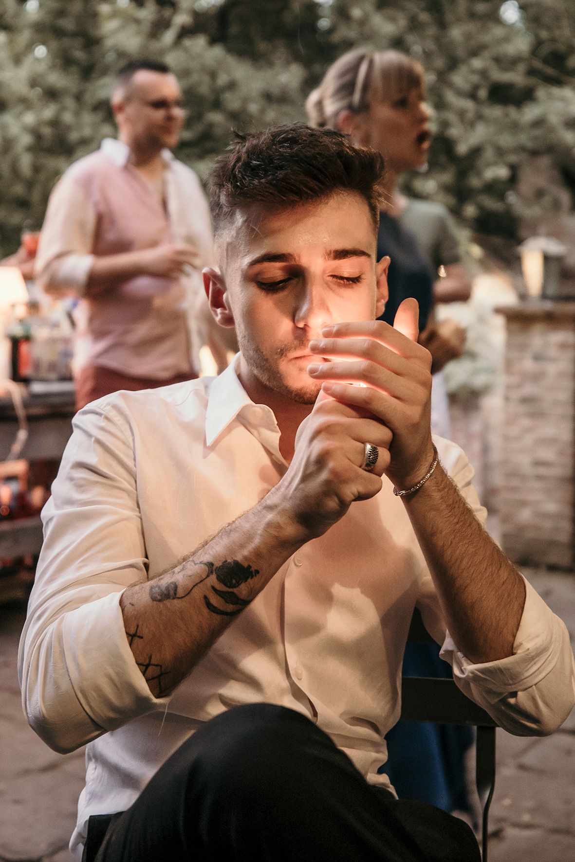 Person in white dress shirt lights a cigarette outdoors while others gather in background during social event.