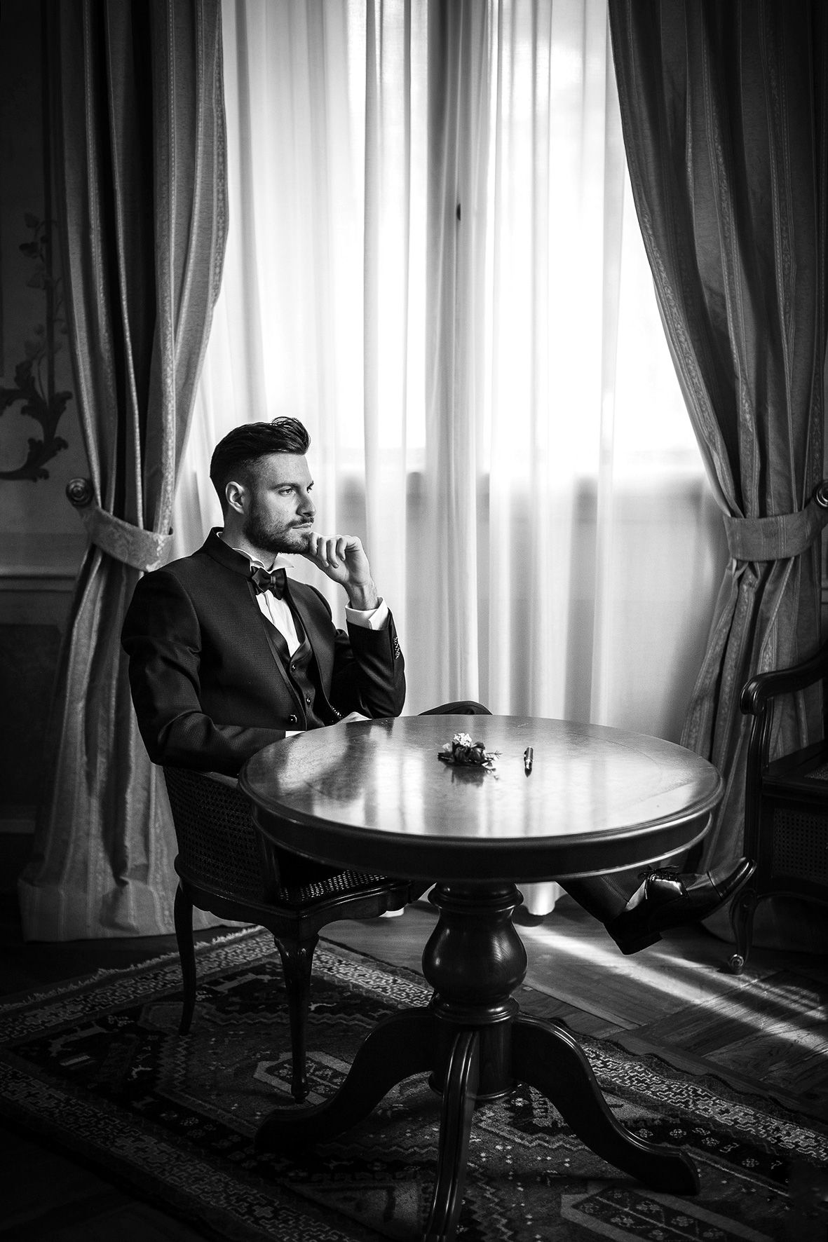 A well-dressed person in a tuxedo sits contemplatively at a round table near elegant curtained windows in black and white.