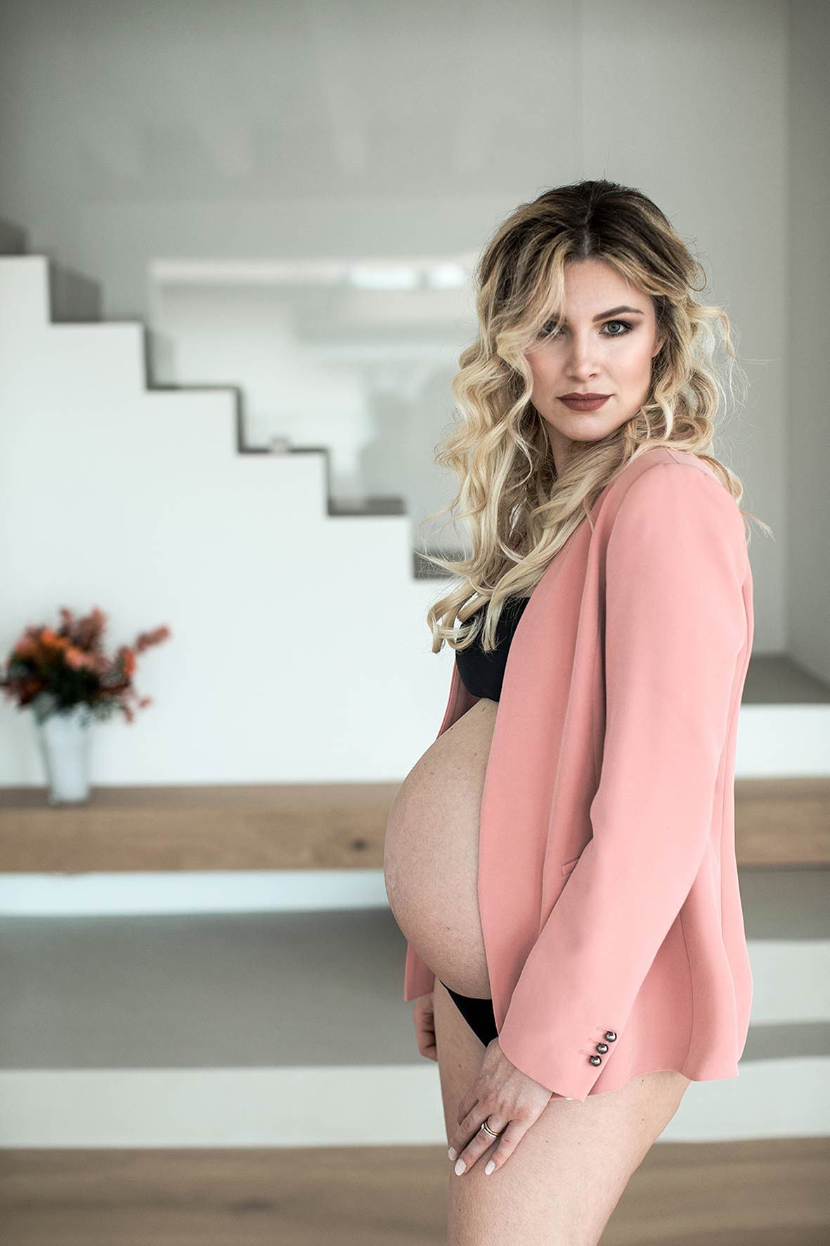 Pregnant woman in black lingerie and pink blazer poses elegantly beside white stairs in a modern interior setting.
