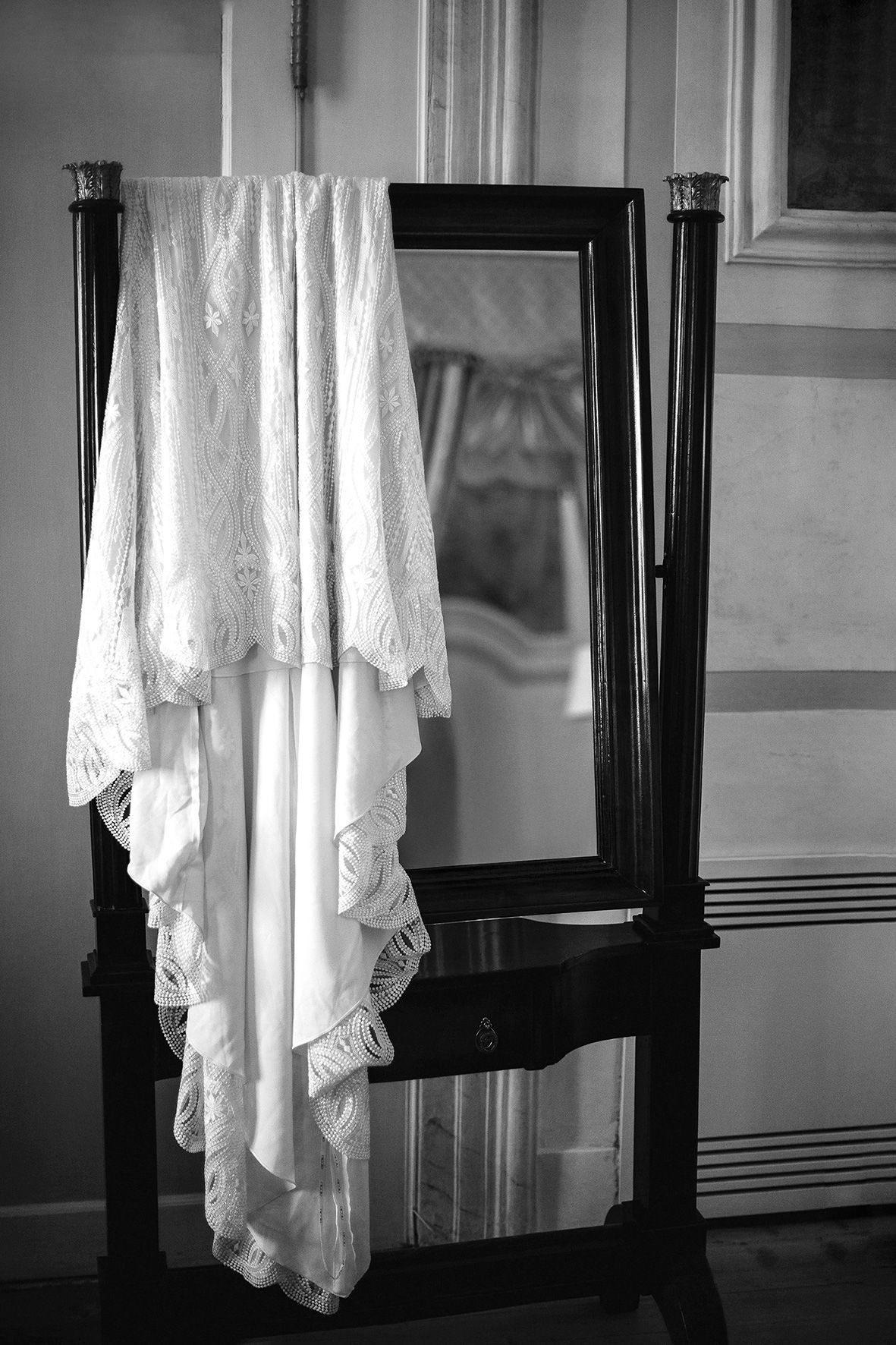 A vintage white lace dress hangs elegantly from a dark wooden mirror stand in a monochrome composition.