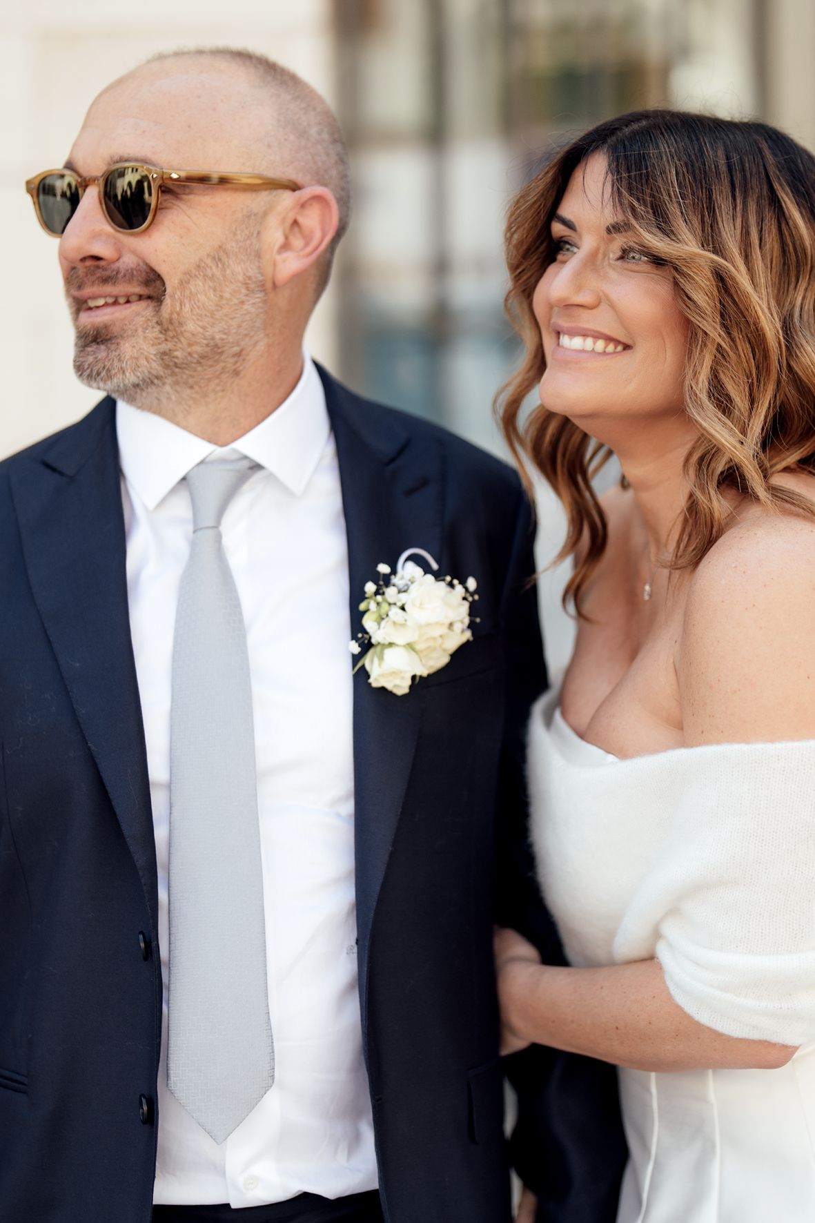 A couple smiles together on their wedding day, with the groom in a navy suit and sunglasses and bride in white dress.