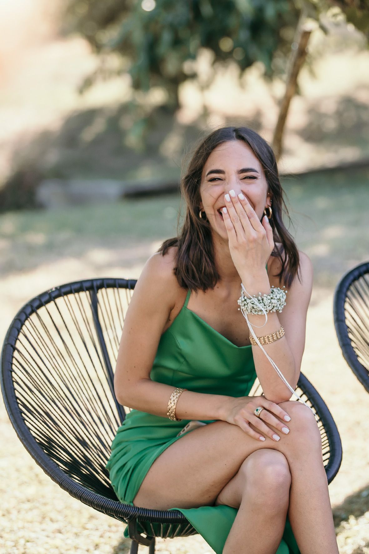 A person in a bright green dress laughs joyfully while sitting in a modern black woven patio chair outdoors.