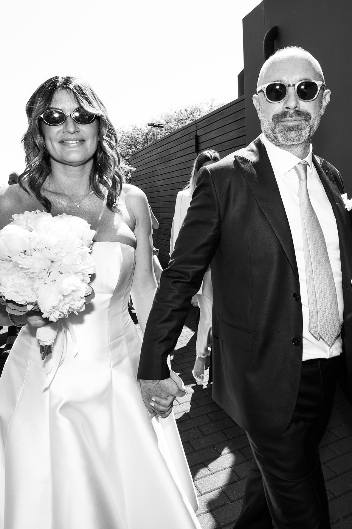 Black and white photo of a bride and groom wearing sunglasses and holding hands on their wedding day.