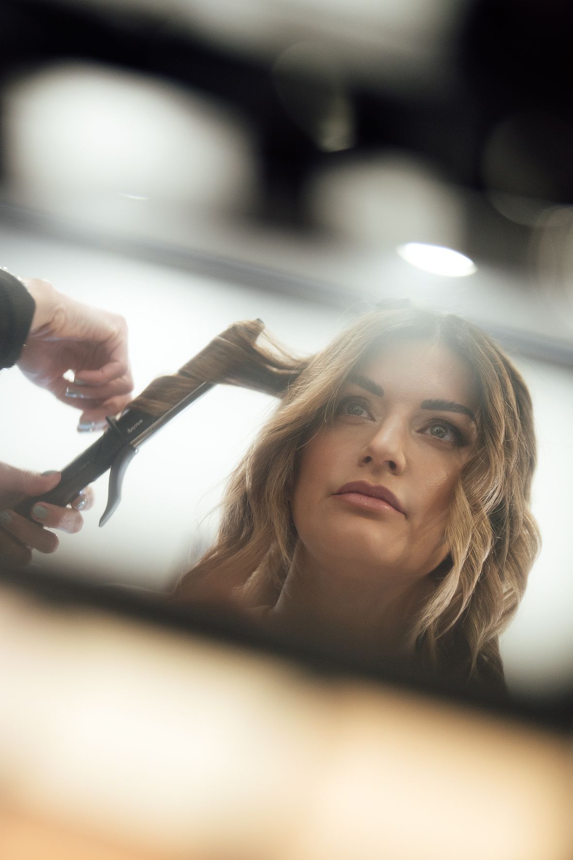 A reflection in a mirror shows a hairstylist working on styling wavy blonde hair in a salon.