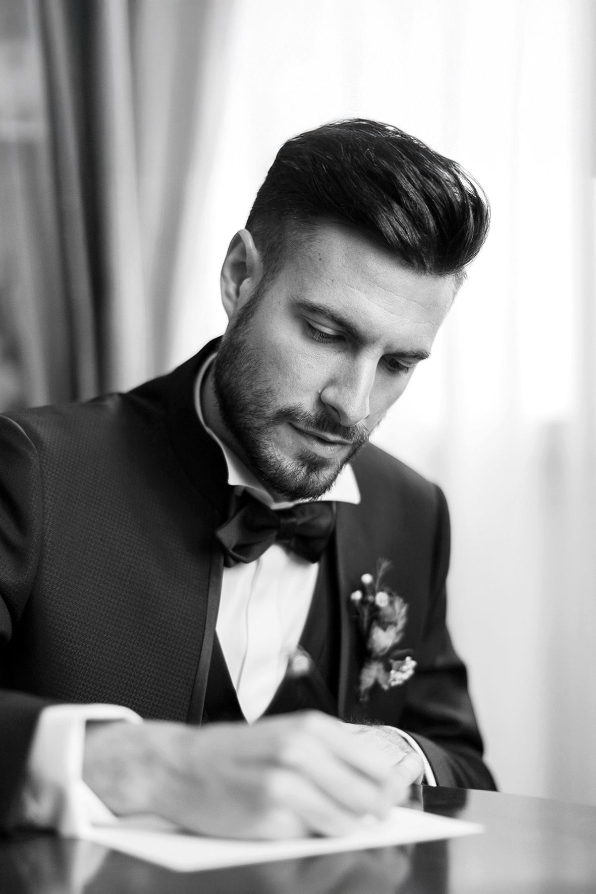 Black and white portrait of a groom in tuxedo and bow tie writing at a desk.
