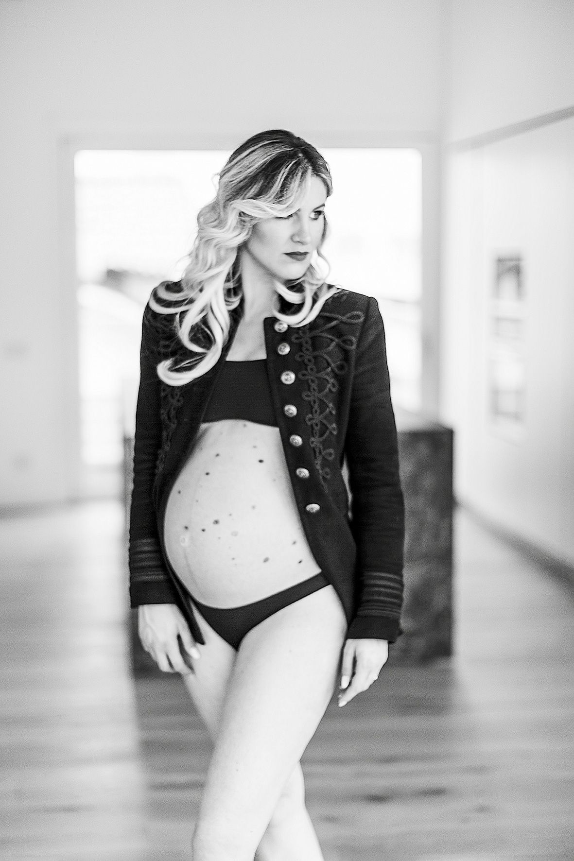 Black and white maternity photo showing expecting mother in military-style jacket and underwear in natural light setting.