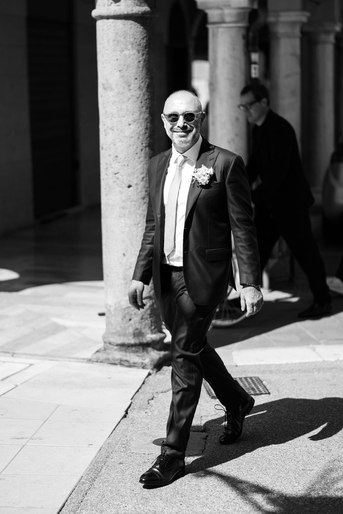 A well-dressed person in a dark suit and sunglasses walks confidently through a columned hallway.