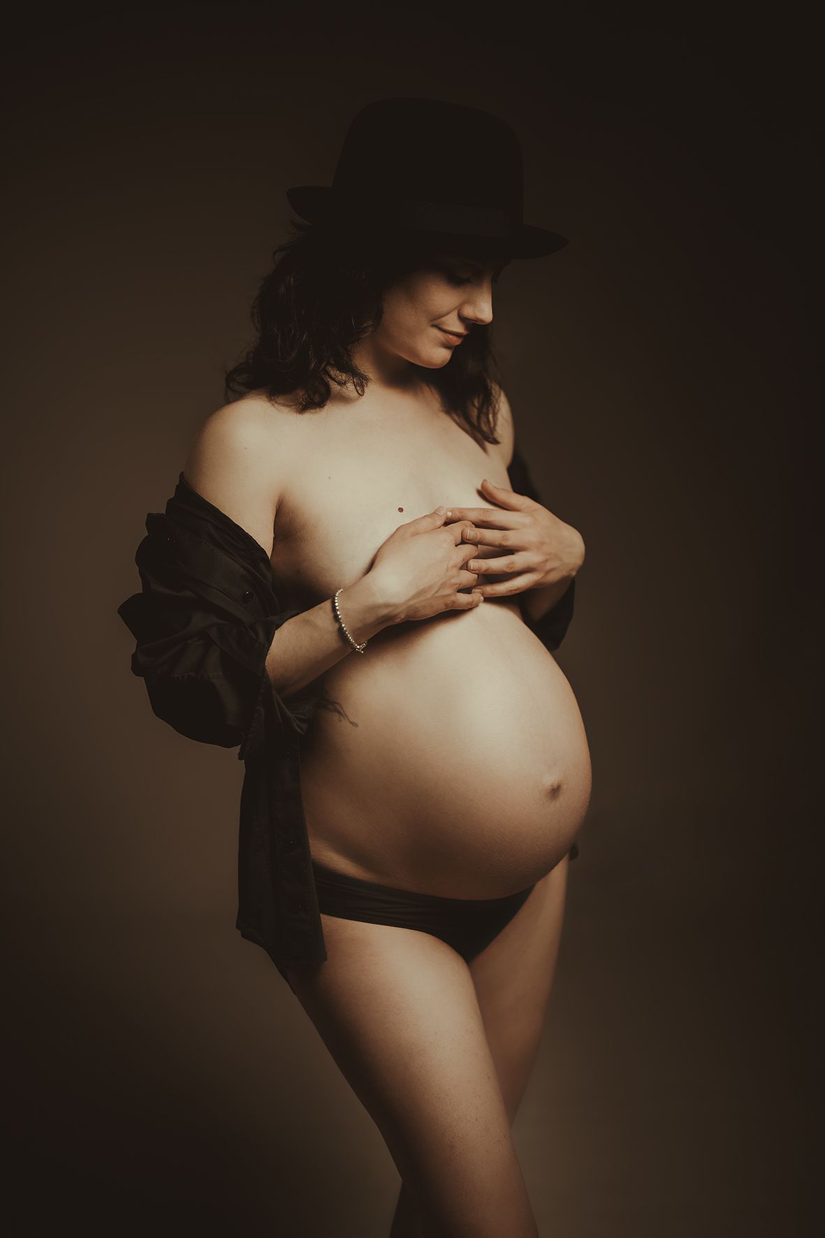 Artistic maternity photo shows silhouette of pregnant figure wearing black hat and open shirt against dark background.