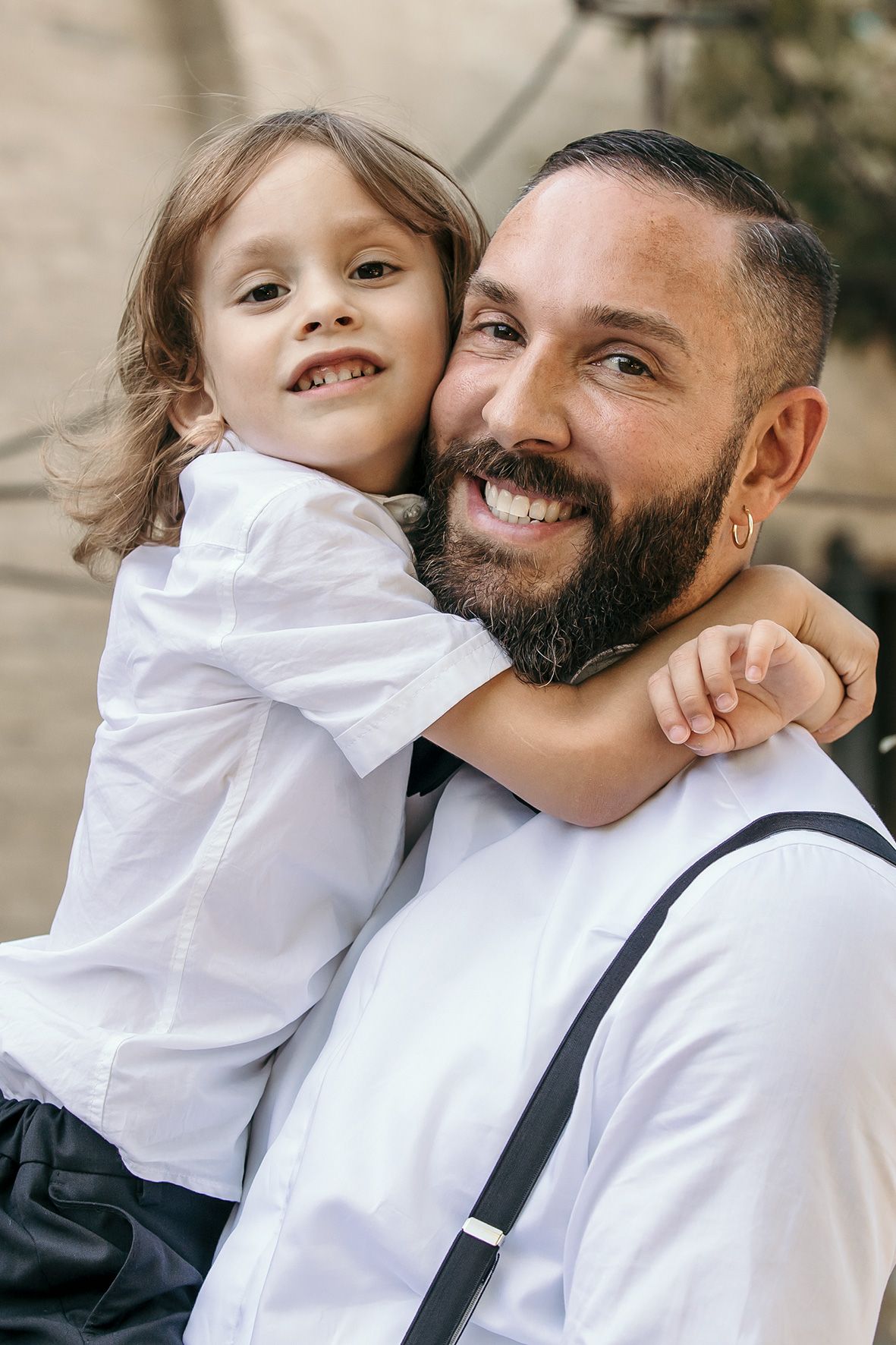 A small child in a white shirt hugging a bearded person wearing suspenders as they both smile warmly at the camera.