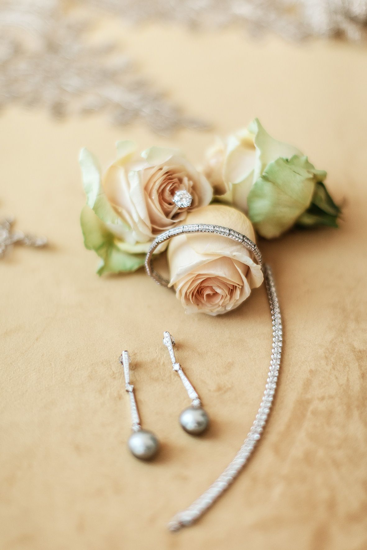 Delicate cream roses paired with elegant silver jewelry including a bracelet and pearl drop earrings on beige background.