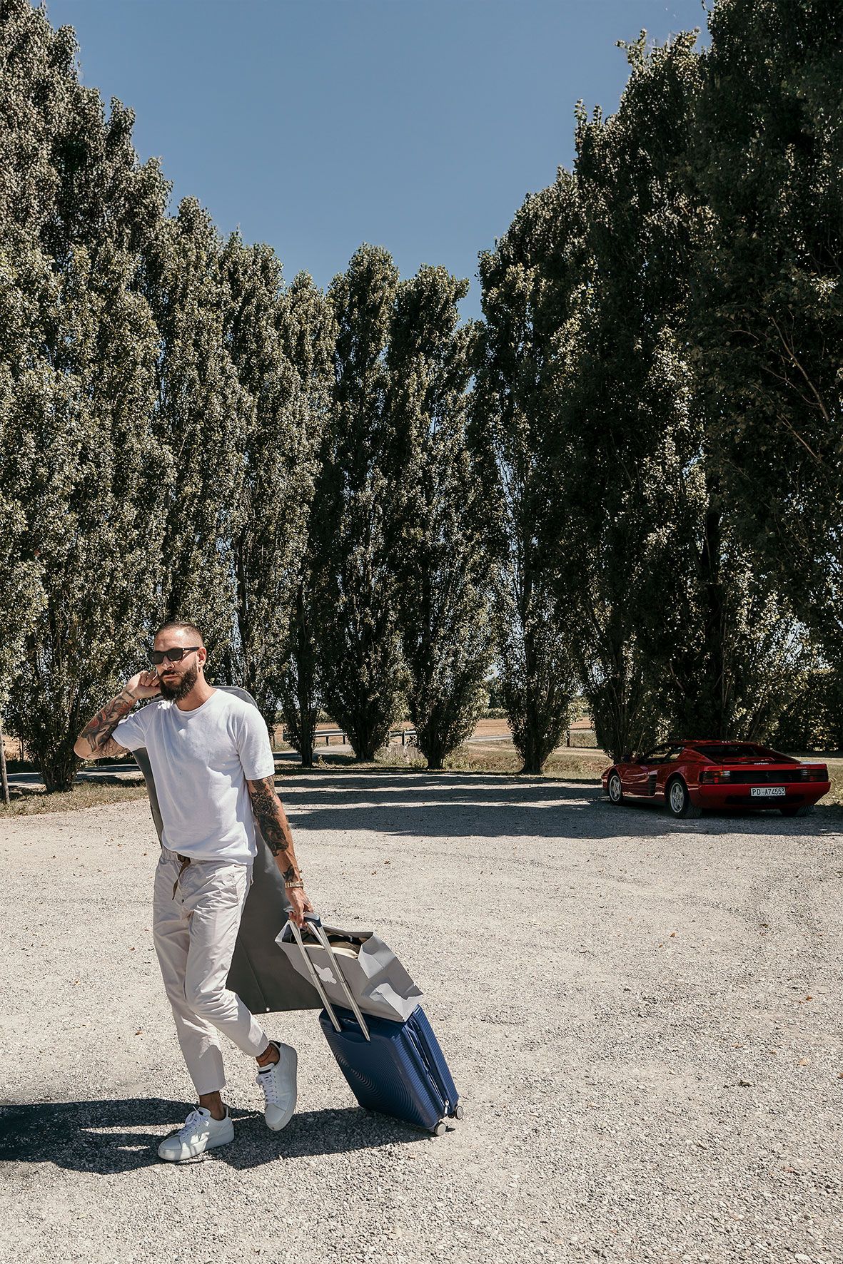 Person with sunglasses wearing white clothing walks with blue suitcase on gravel path lined with tall trees and red car parked.