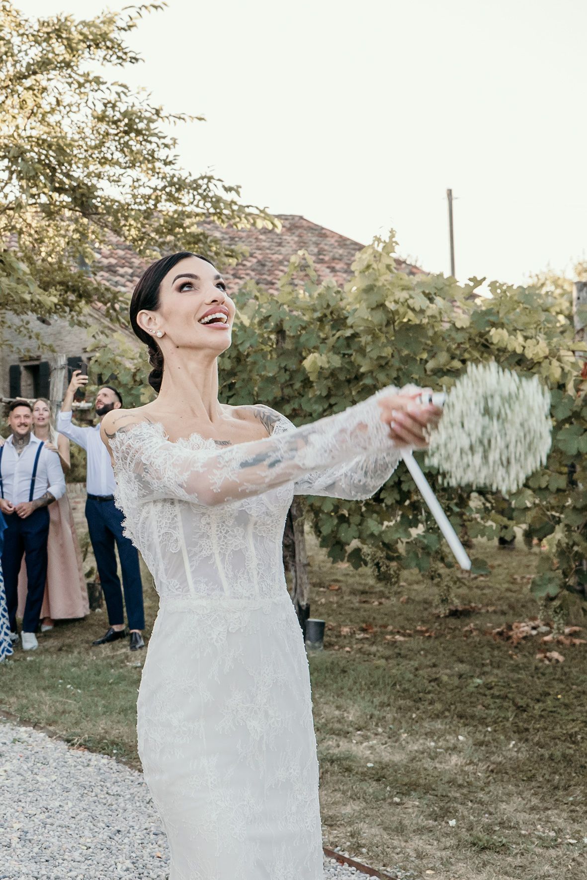 A bride in a fitted lace wedding dress joyfully tosses confetti in an outdoor garden setting at dusk.