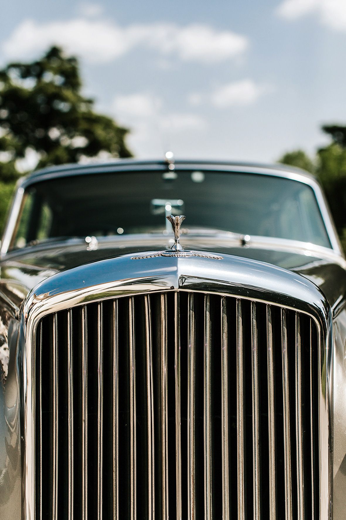 Close-up view of a classic Bentley's iconic chrome grille and hood ornament against a blue sky background.