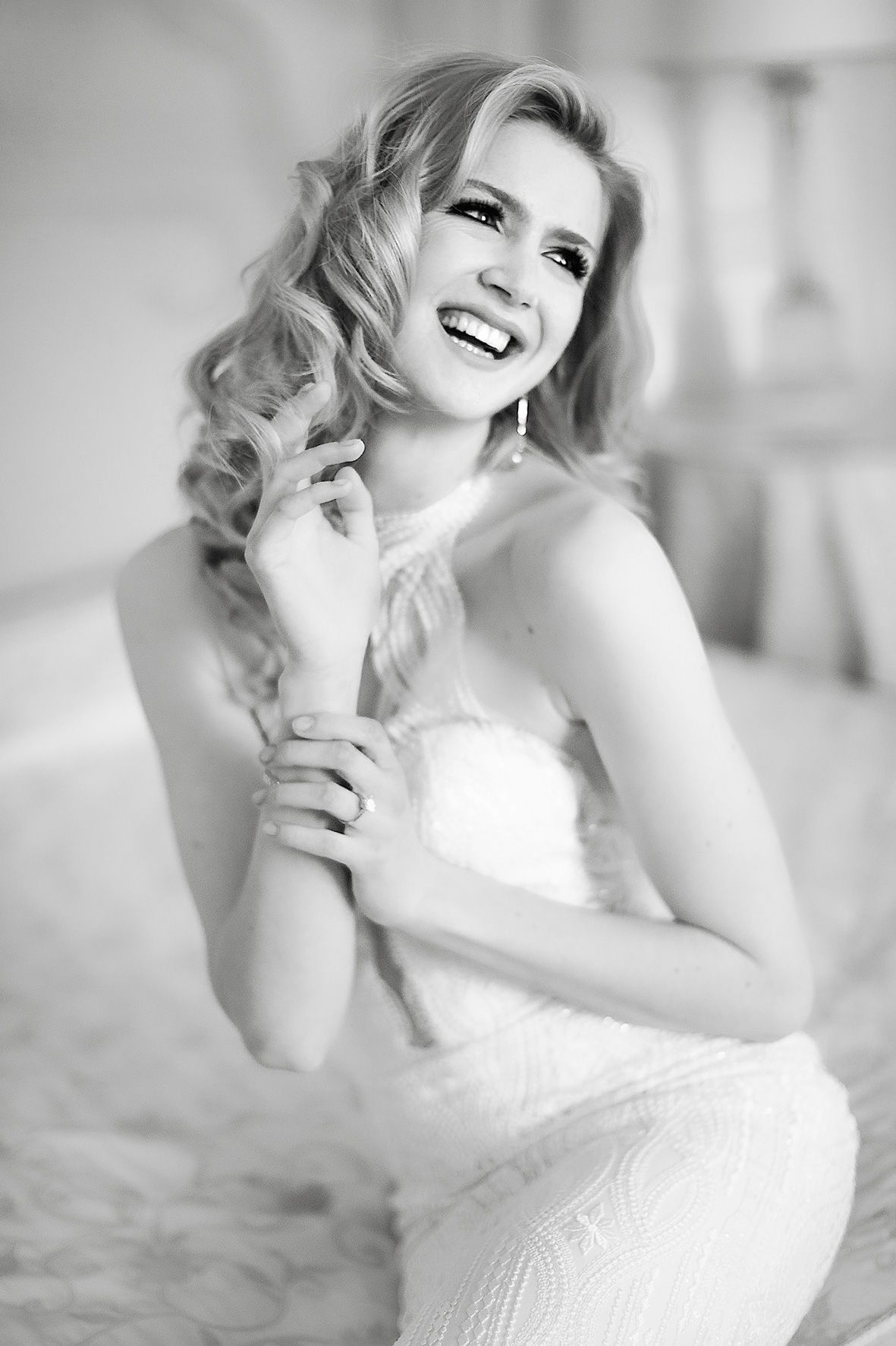 Black and white portrait of a joyful bride wearing a strapless lace wedding dress and wavy blonde hair.