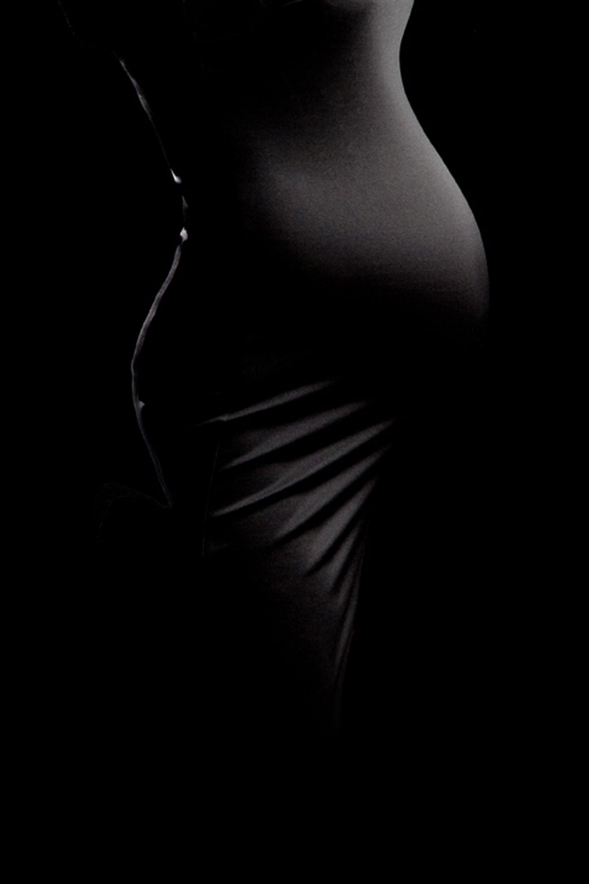 Black and white artistic silhouette of a pregnant belly in dramatic side lighting showing gentle curves and fabric draping.