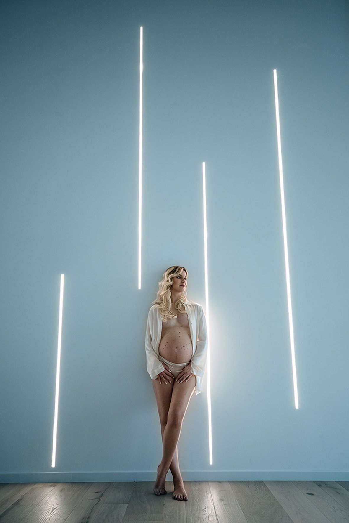 Figure in white sweater and shorts stands against blue wall with vertical fluorescent light tubes creating dramatic ambiance.