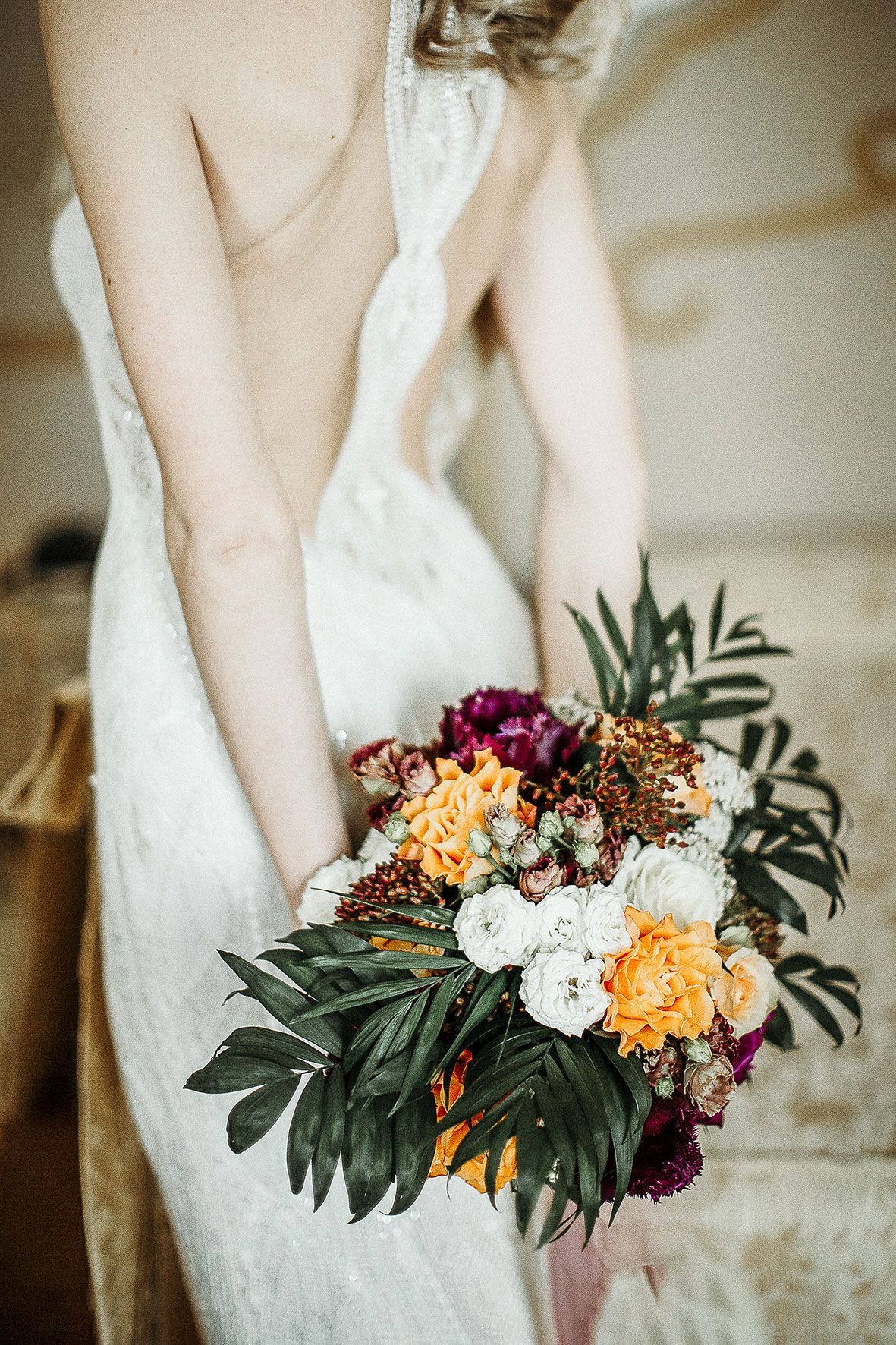 A wedding bouquet with burgundy, orange and white flowers mixed with tropical leaves held against a white lace dress.
