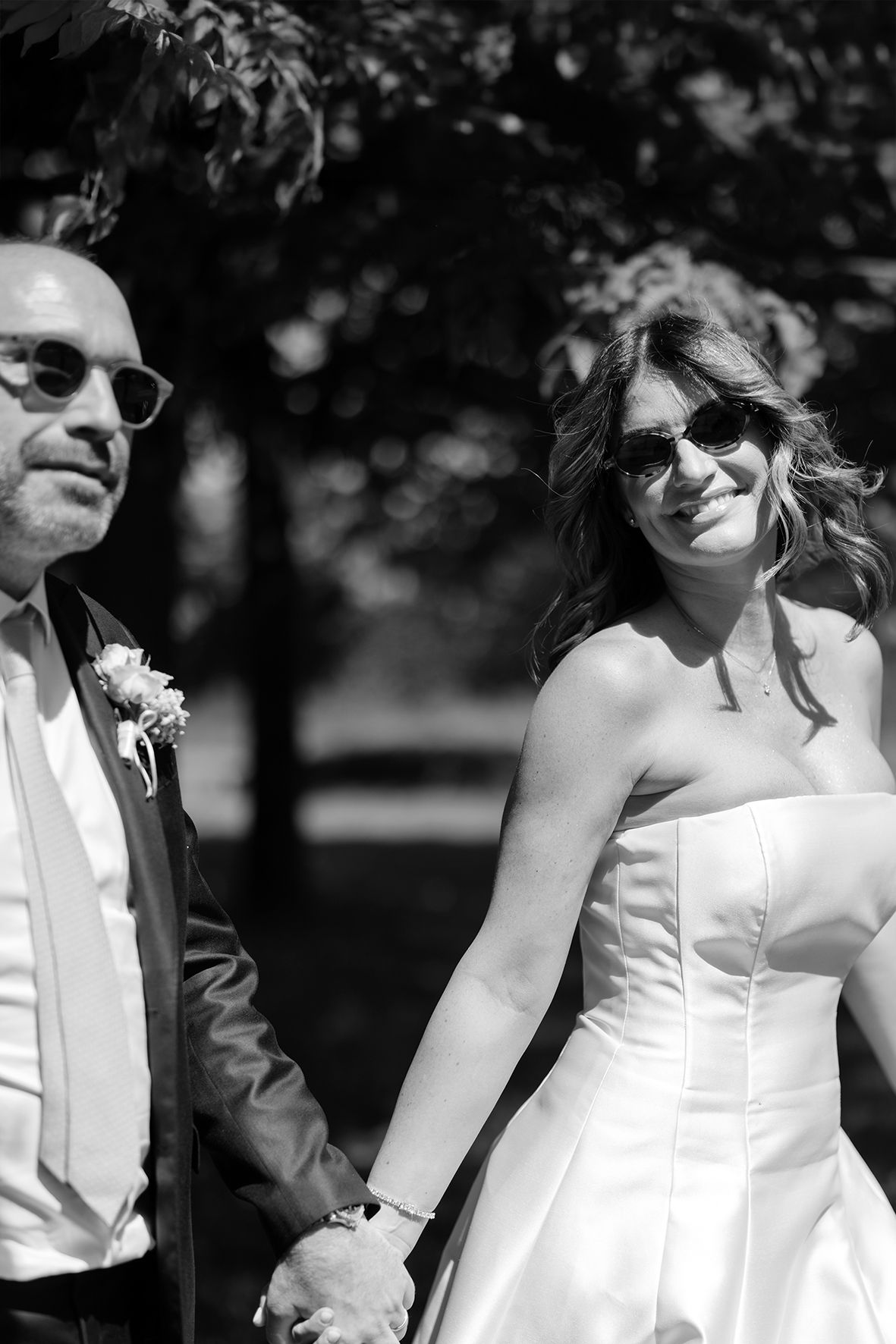 Black and white photo of a newlywed couple outdoors in formal wedding attire holding hands and wearing sunglasses.