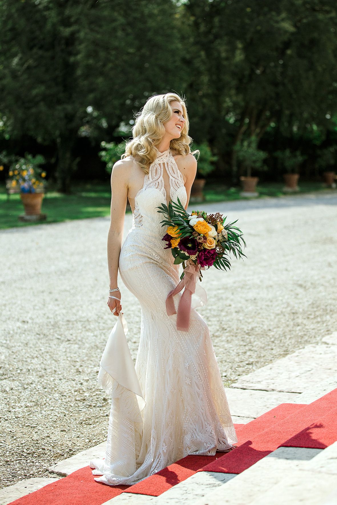 Elegant wedding dress silhouette on red carpet path in garden setting with vibrant floral bouquet.