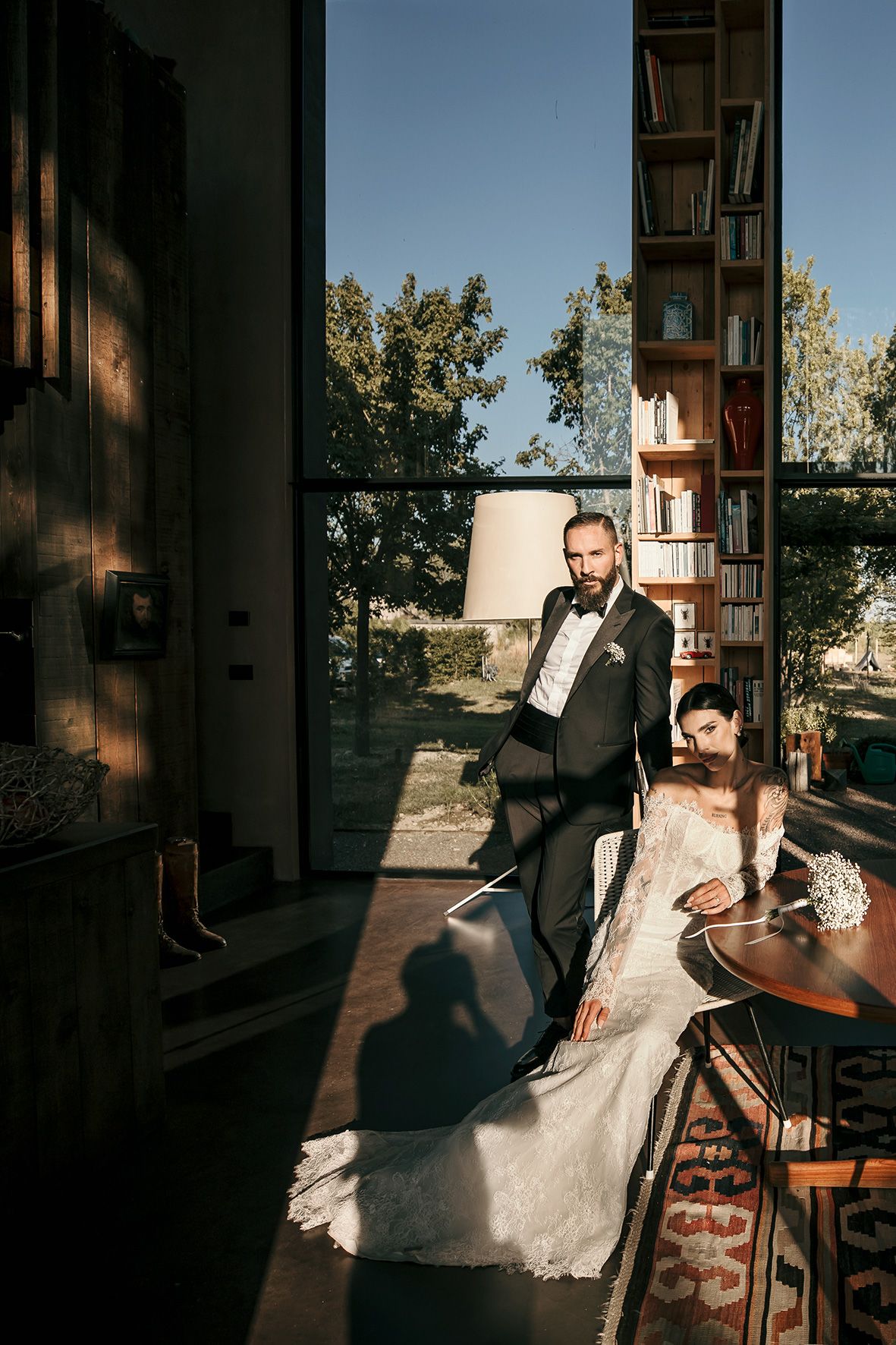 A couple poses in formal attire by a tall bookshelf near large windows with natural sunlight creating dramatic shadows.