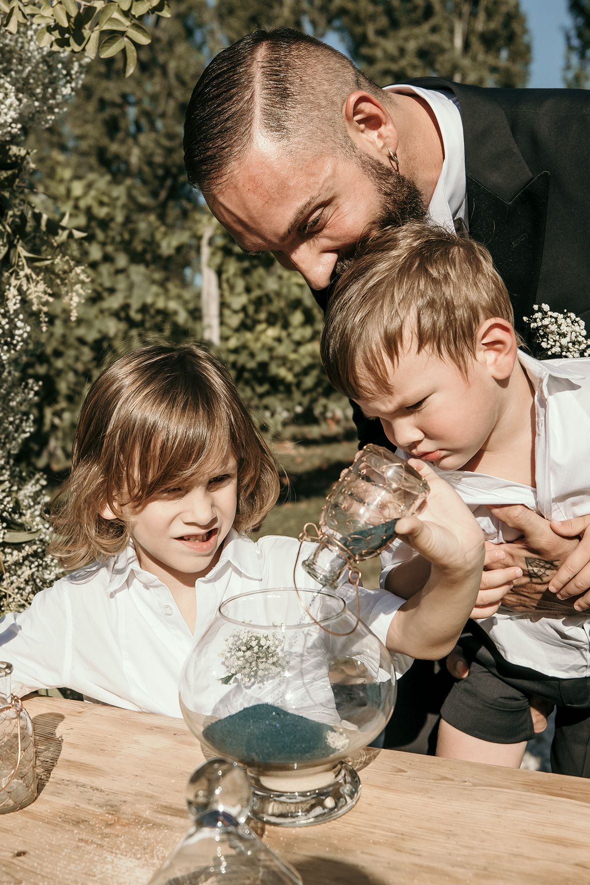 A parent and two children gather around a table outdoors examining glass containers in natural sunlight.